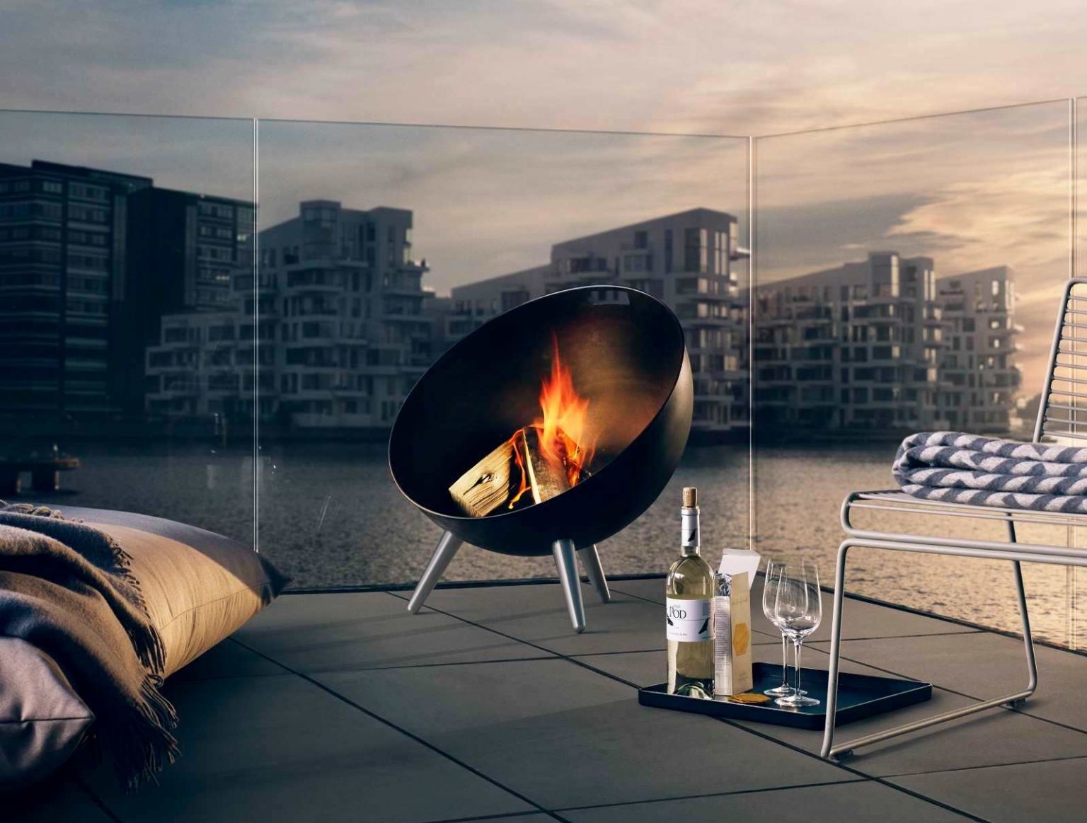 Moderne Feuerstelle für den Außenbereich auf einer Terrasse mit Blick auf das Wasser und Stadtgebäude.