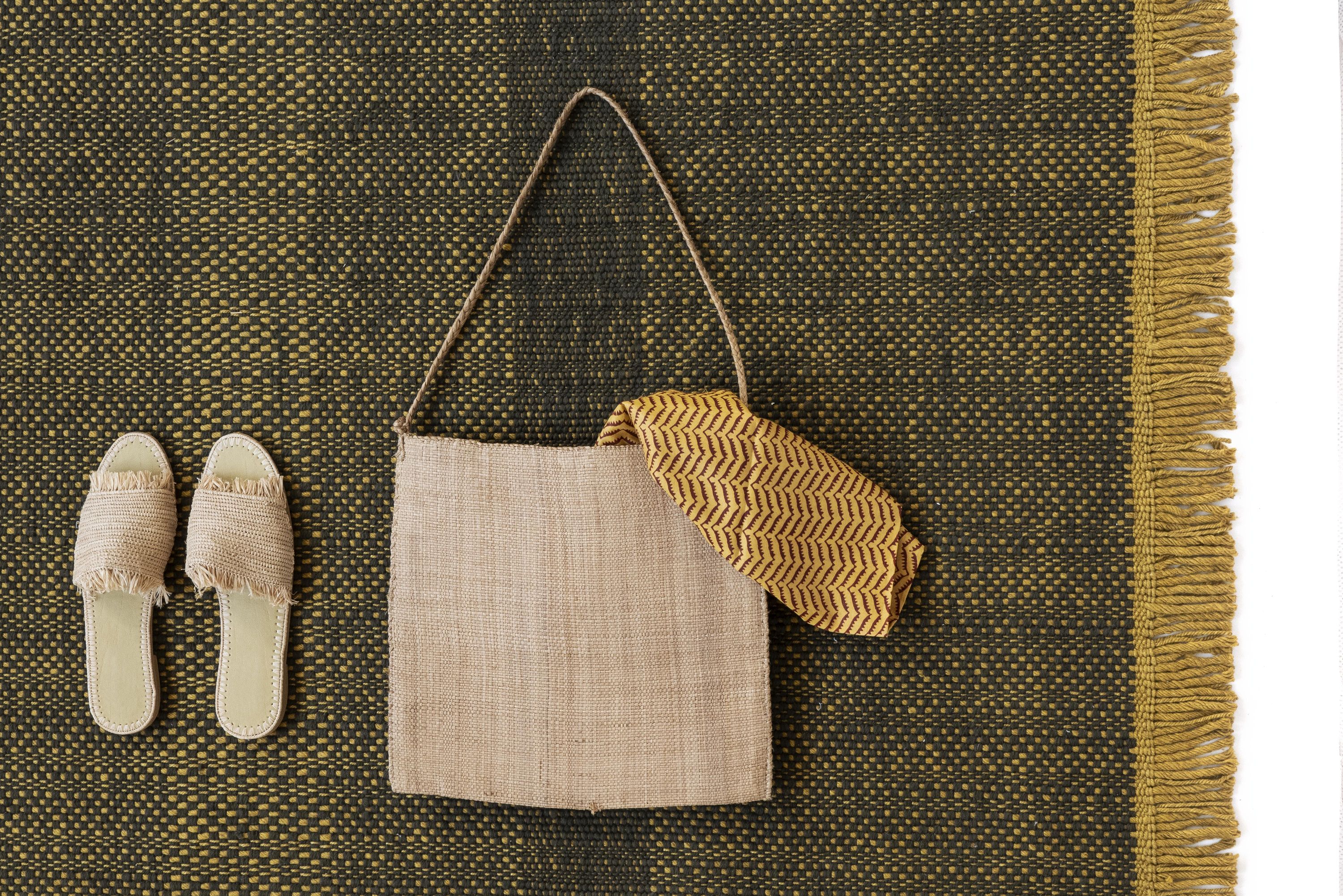 Detailaufnahme des Tres Outdoor Texture Teppichs in Senf mit Tasche und Sandalen, Textur und Fransen sichtbar.