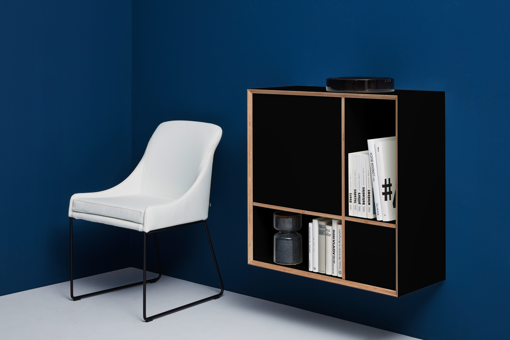 Vertiko Ply Regal von Müller Möbelwerkstätten in Schwarz mit Büchern und Dekoration, neben weißem Stuhl.