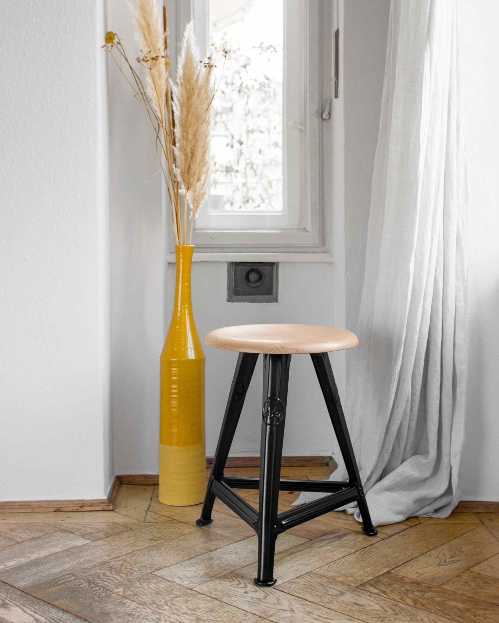 Schemel Model I: Hocker mit Holzplatte und schwarzem Metallgestell vor Fenster.