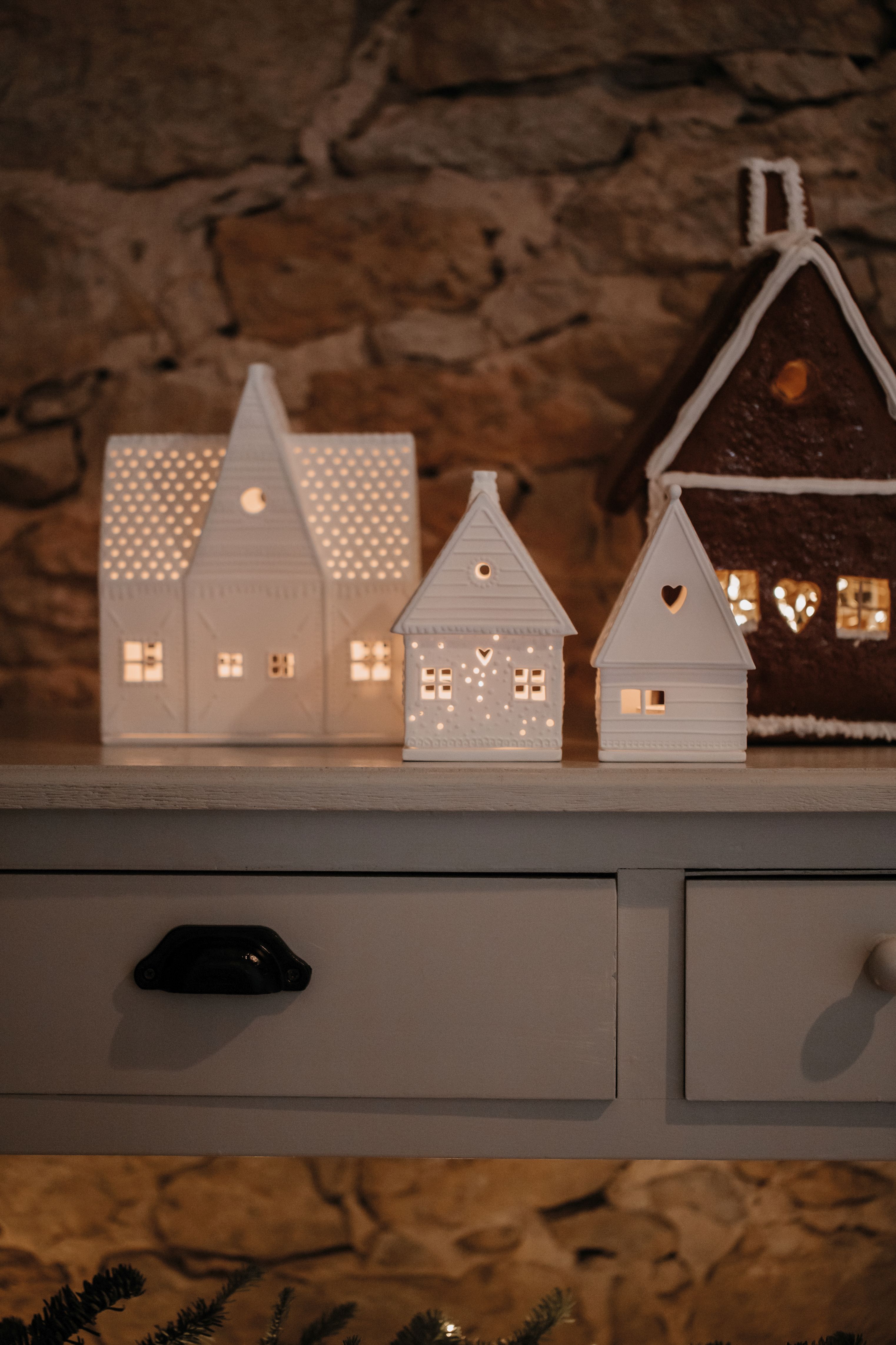 Drei beleuchtete Lebkuchenhäuser, darunter ein Räder Lichthaus, auf einer Kommode vor einer Steinwand.