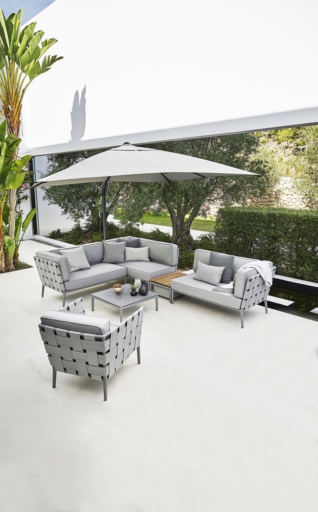 Weißer Hyde Luxe Sonnenschirm von Cane-Line mit Fuß über grauer Lounge-Garnitur im Garten.