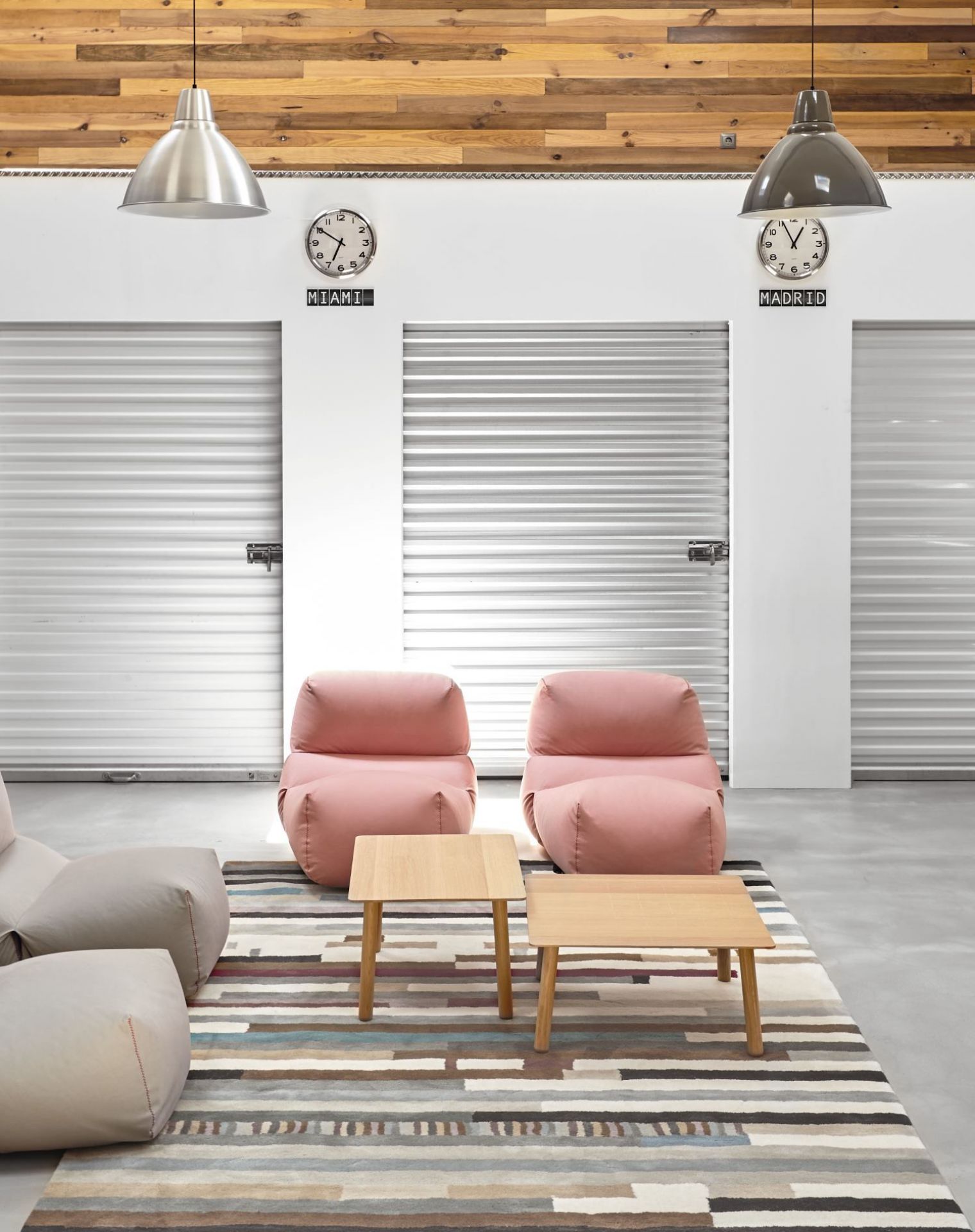 Wohnzimmer mit rosa Sessel, beigem Sofa, Holztischen und gestreiftem Teppich vor Lagerraumtüren.