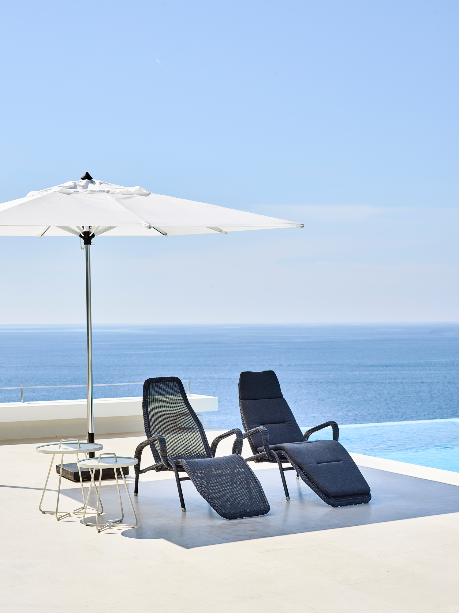 Zwei Cane-Line Sonnenliegen mit Sonnenschirm und Beistelltischen am Pool mit Meerblick.