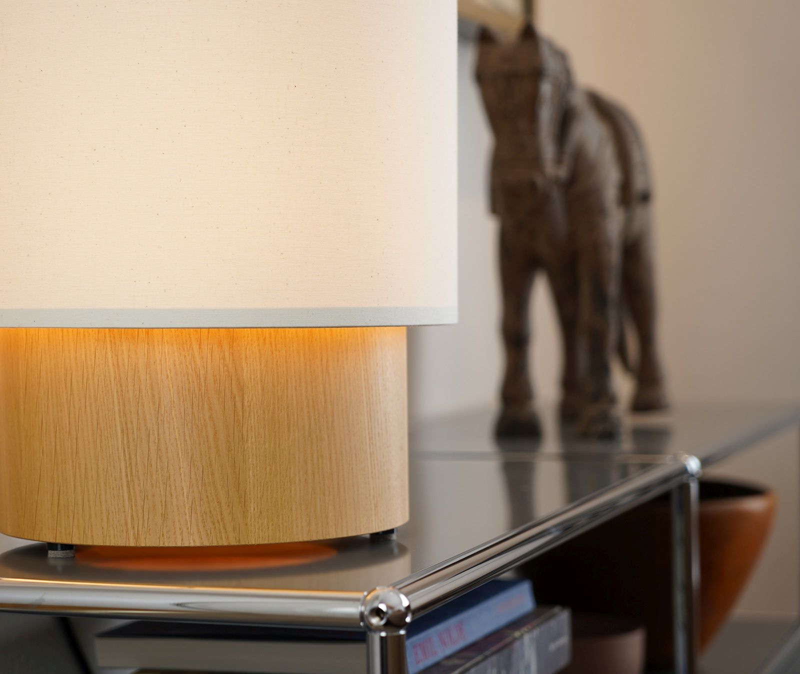 Hiro Tischleuchte von Maigrau mit hellem Lampenschirm und Holzfuß auf einem Regal.