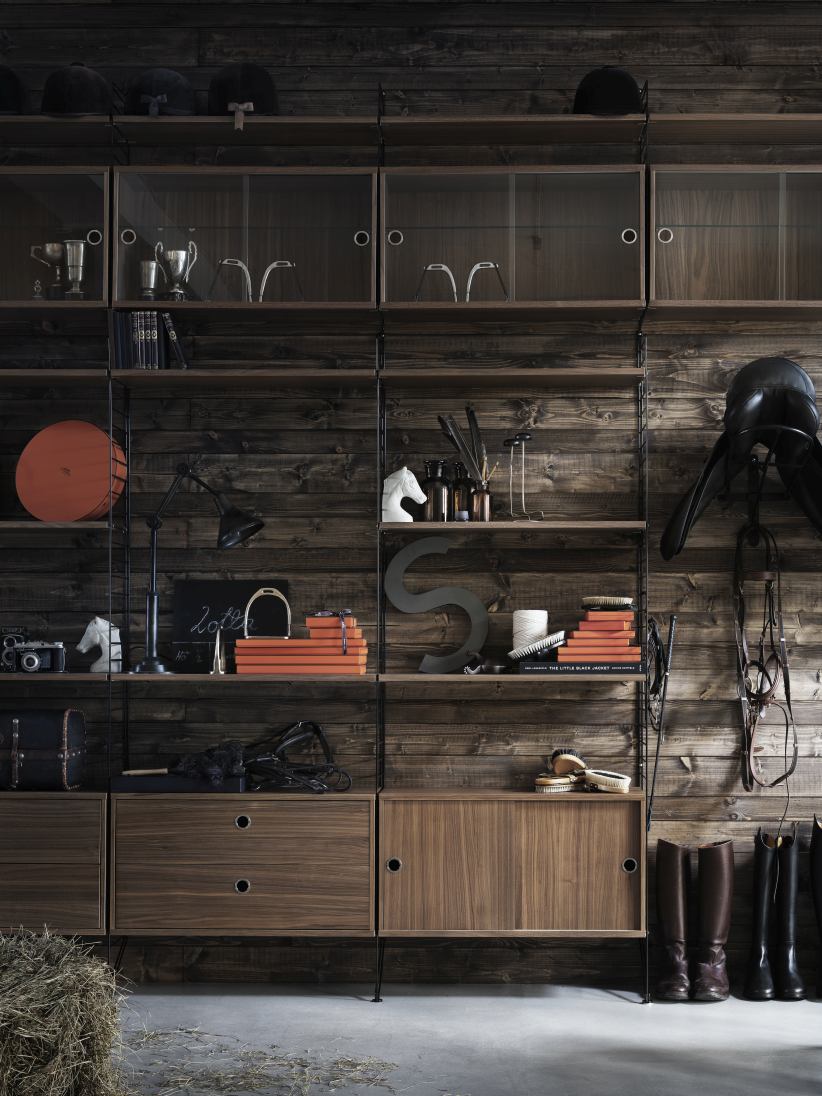 Wohnzimmer KONFIGURATIONEN L8 String Furniture Regalsystem aus Holz mit Reitsport-Accessoires vor rustikaler Holzwand.