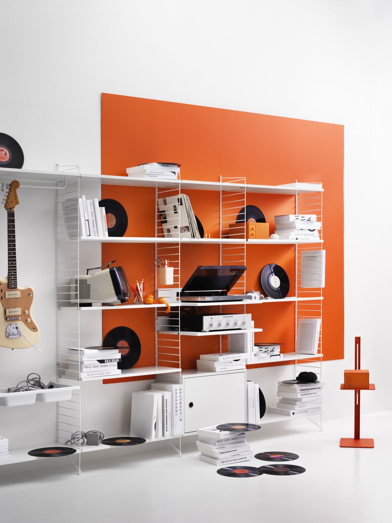 Weißes String Regalsystem mit Schallplatten, Büchern und Gitarre vor oranger Wand. Wohnzimmer Regale.