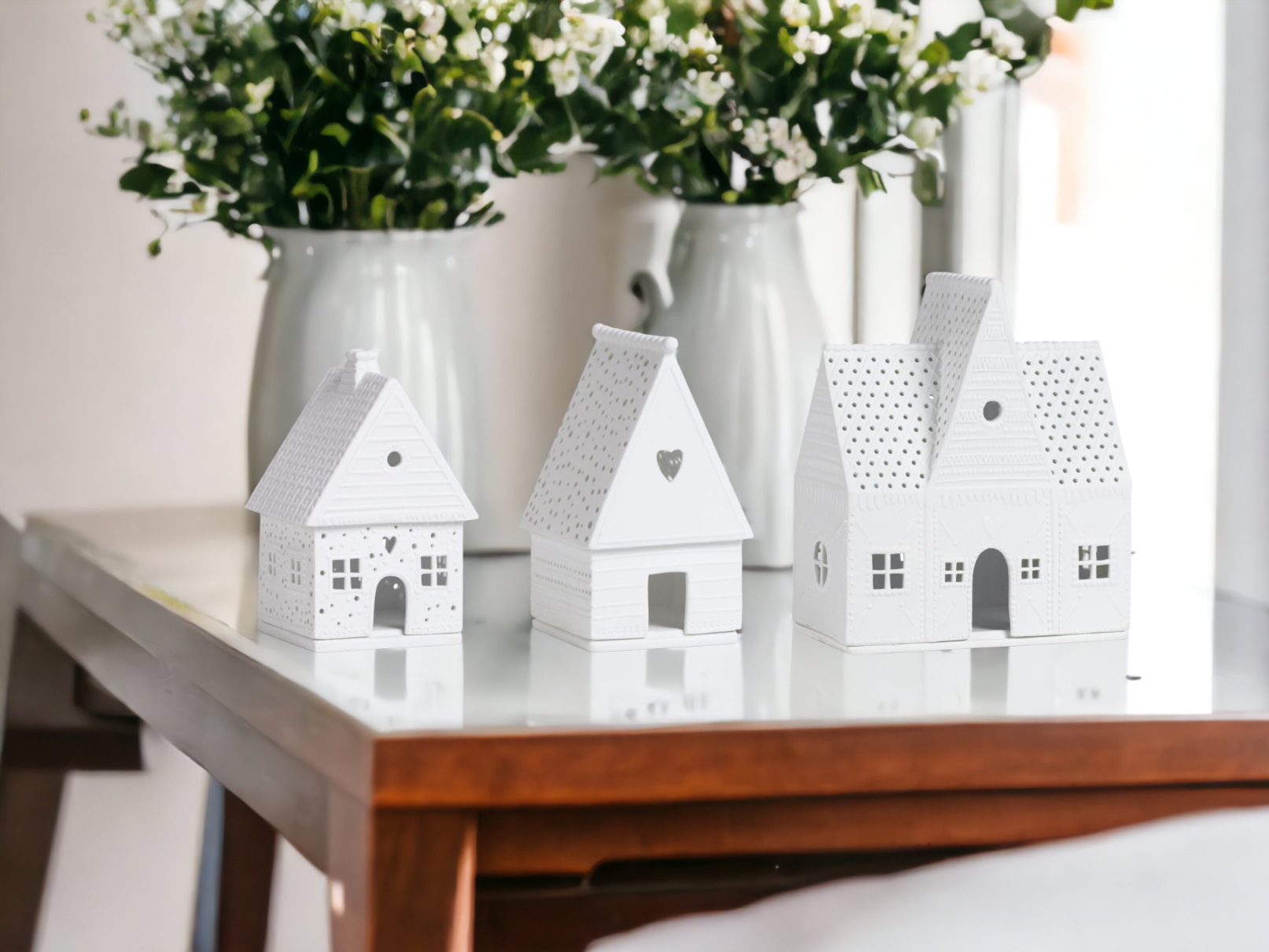 Drei weiße Keramik-Häuser als Dekoration auf einem Tisch, mit Blumenvasen im Hintergrund.