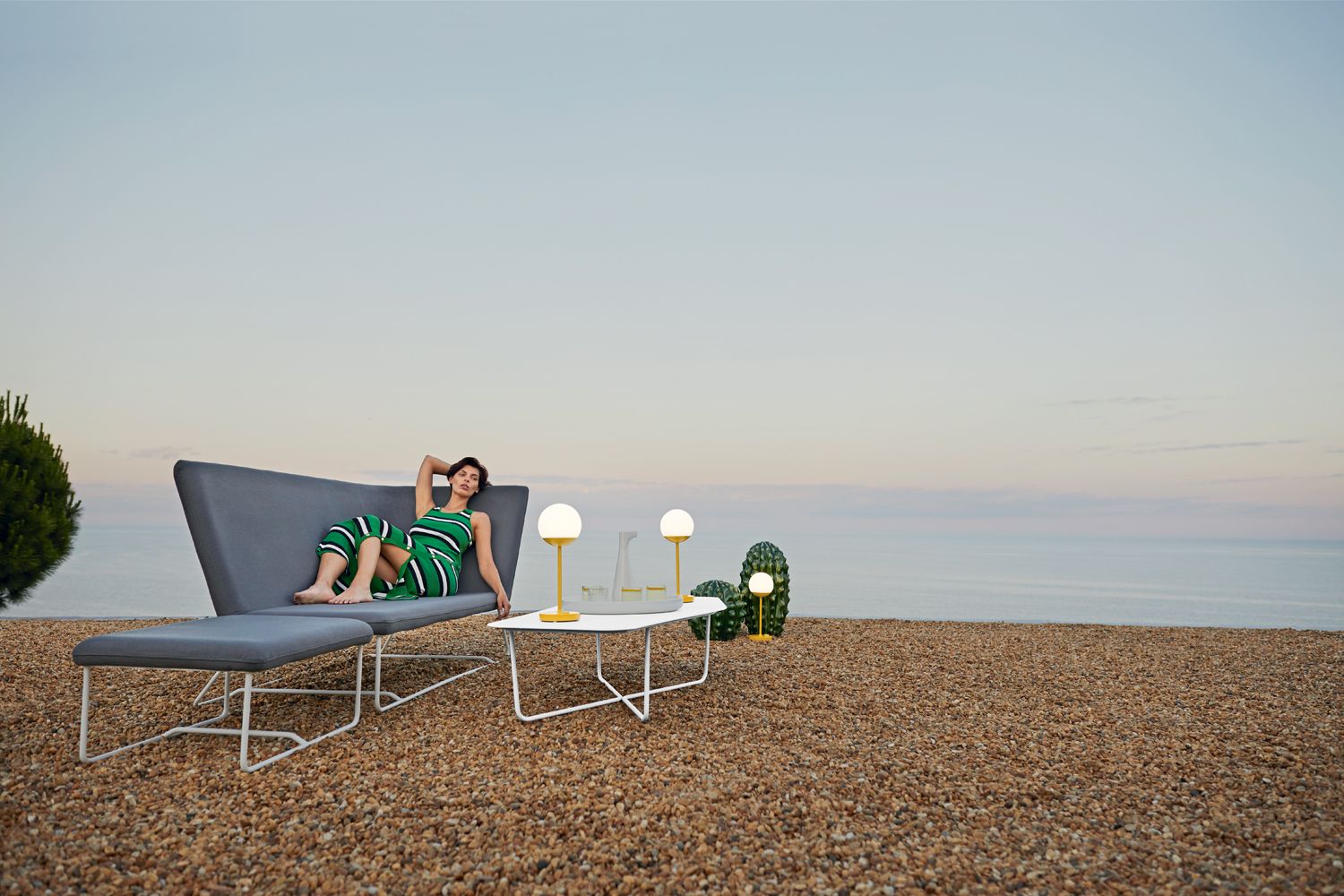 Frau entspannt auf Fermob Ultrasofa Outdoor-Lounge mit niedrigem Tisch und Leuchten am Meer.