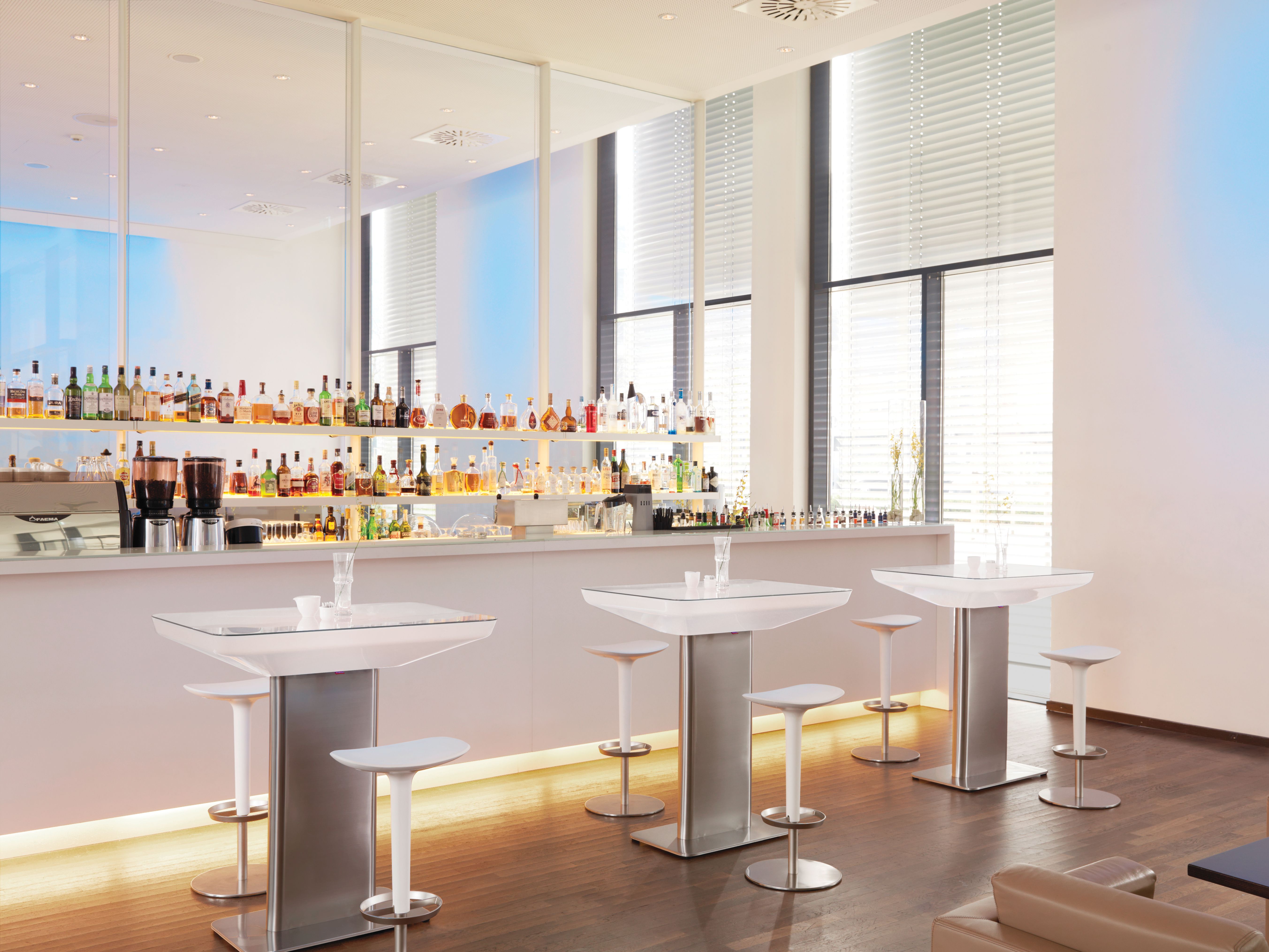 Heller Raum mit Moree Stehtischen und Barhockern vor einer Bar mit Spirituosen.