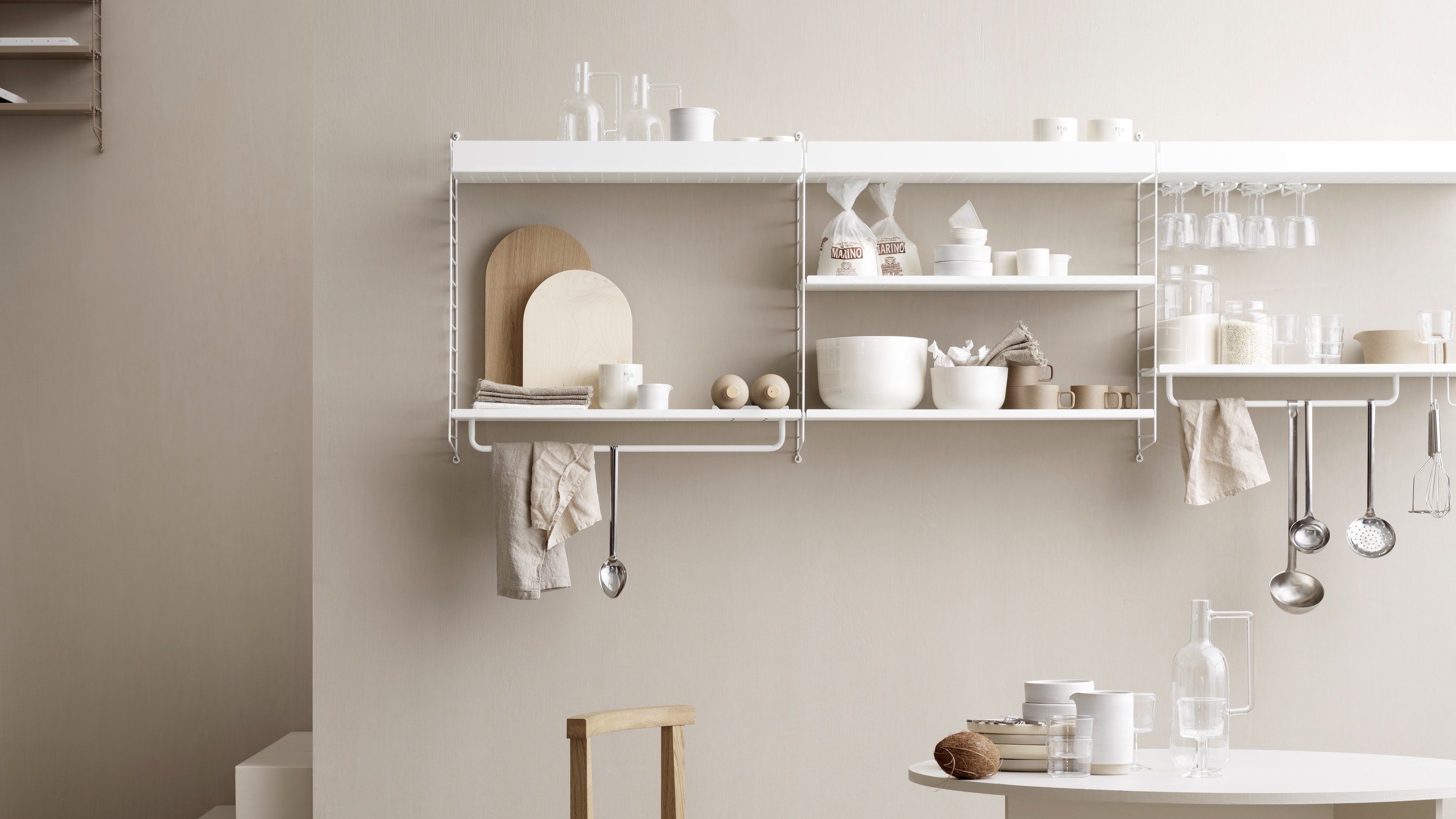 Weißes String Regal mit Küchenutensilien, Geschirr und Lebensmitteln vor beiger Wand, minimalistisches Küchendesign.