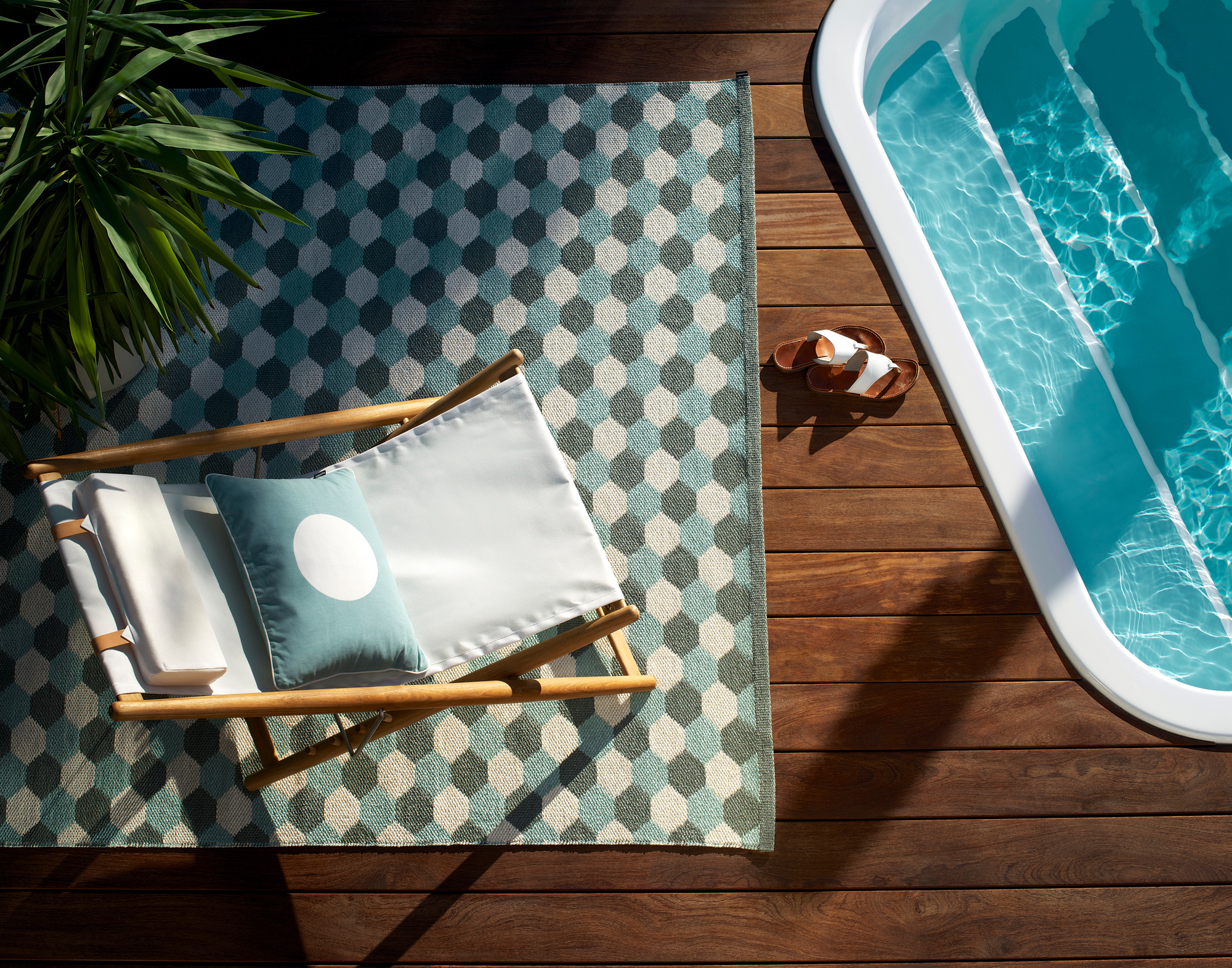 Draufsicht: Pappelina Kunststoffteppich mit geometrischem Muster, Liegestuhl und Pool auf Holzterrasse.