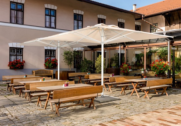 Weishäupl Sonnenschirm: Quadratischer, beiger Teleskopschirm spendet Schatten über Tischen und Bänken im Außenbereich eines Restaurants.