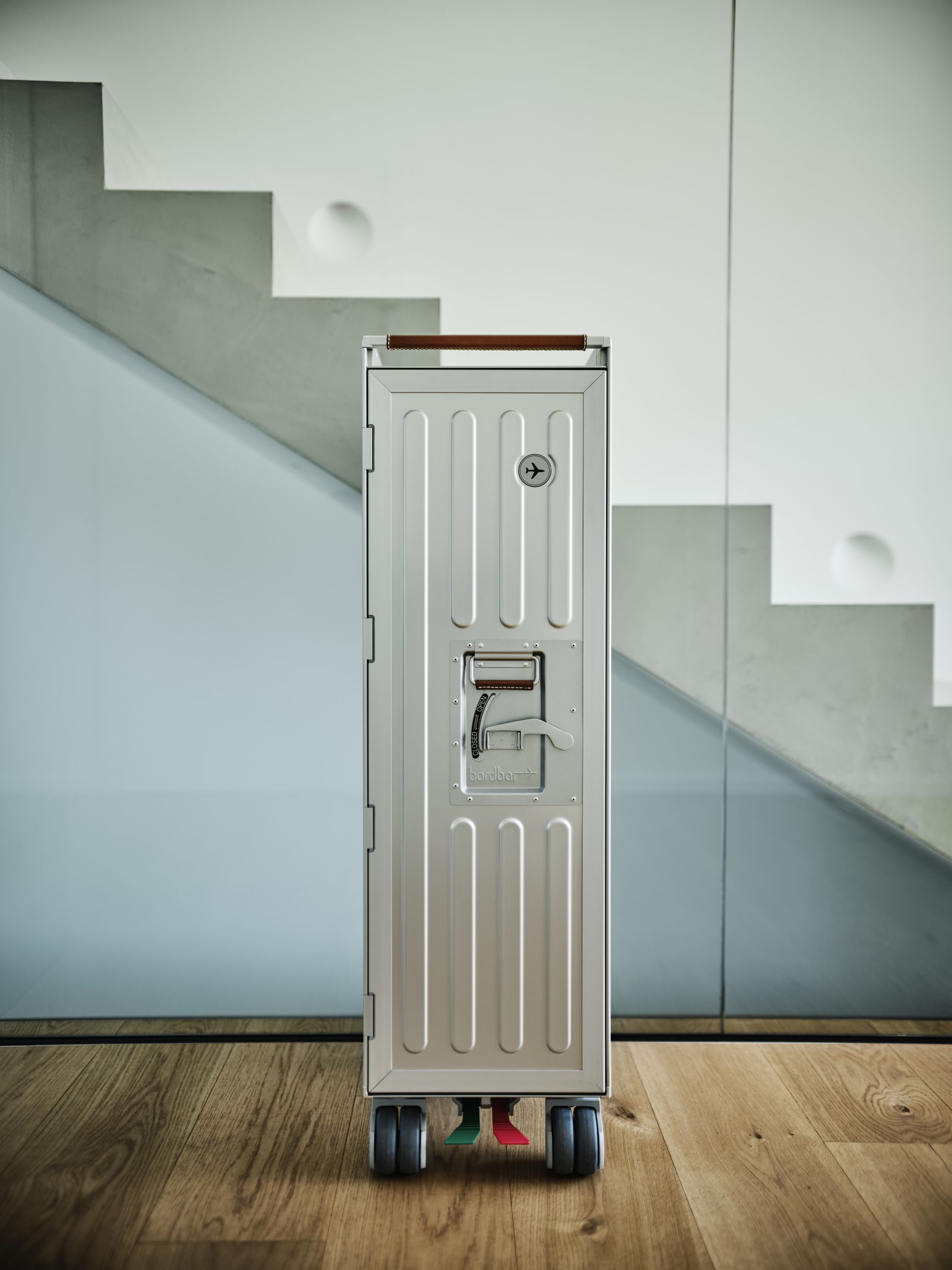 Voyager Flugzeugtrolley Bordbar aus Aluminium mit braunem Ledergriff vor einer modernen Treppe und Holzboden.