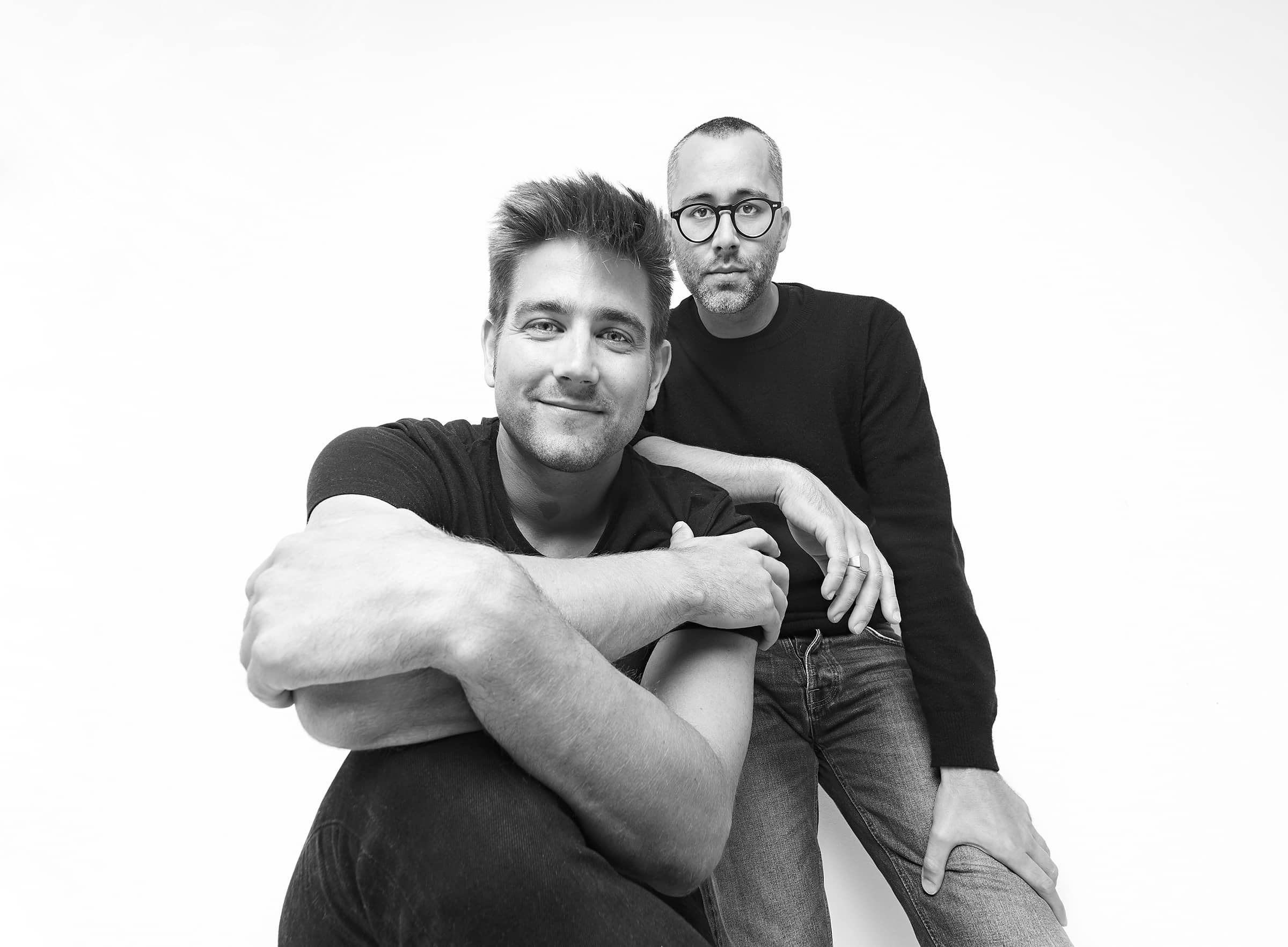 Schwarz-Weiß-Porträt von zwei Männern, lächelnd, mit Brille, in Jeans und Pullover.