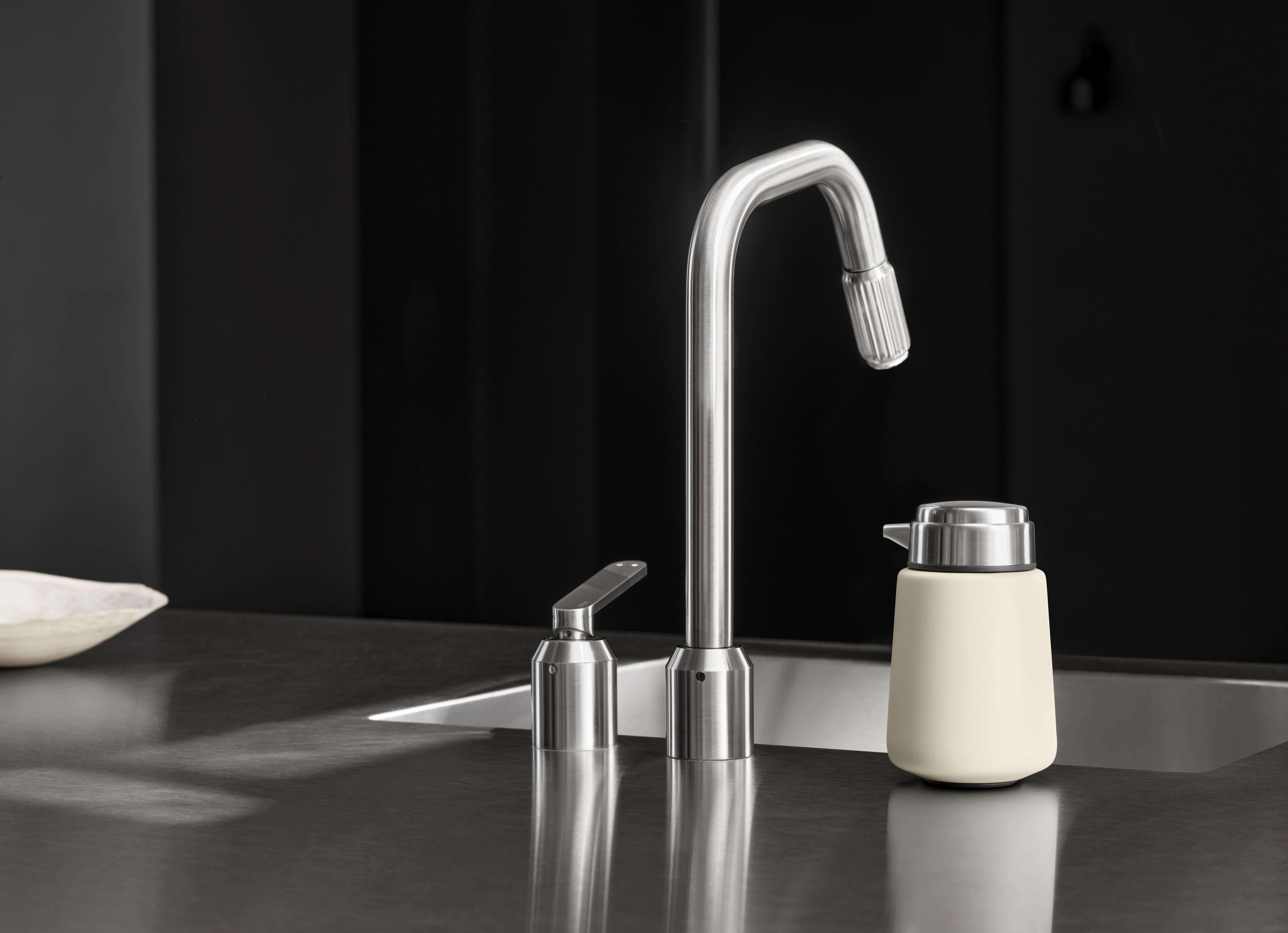 Beiger Seifenspender von Vipp neben einer modernen Küchenarmatur auf einer dunklen Arbeitsplatte.