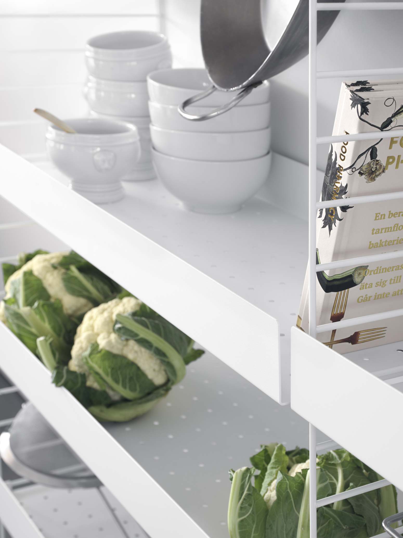 Detailaufnahme eines weißen String Regalsystems mit Geschirr, Kochbuch und Gemüse.