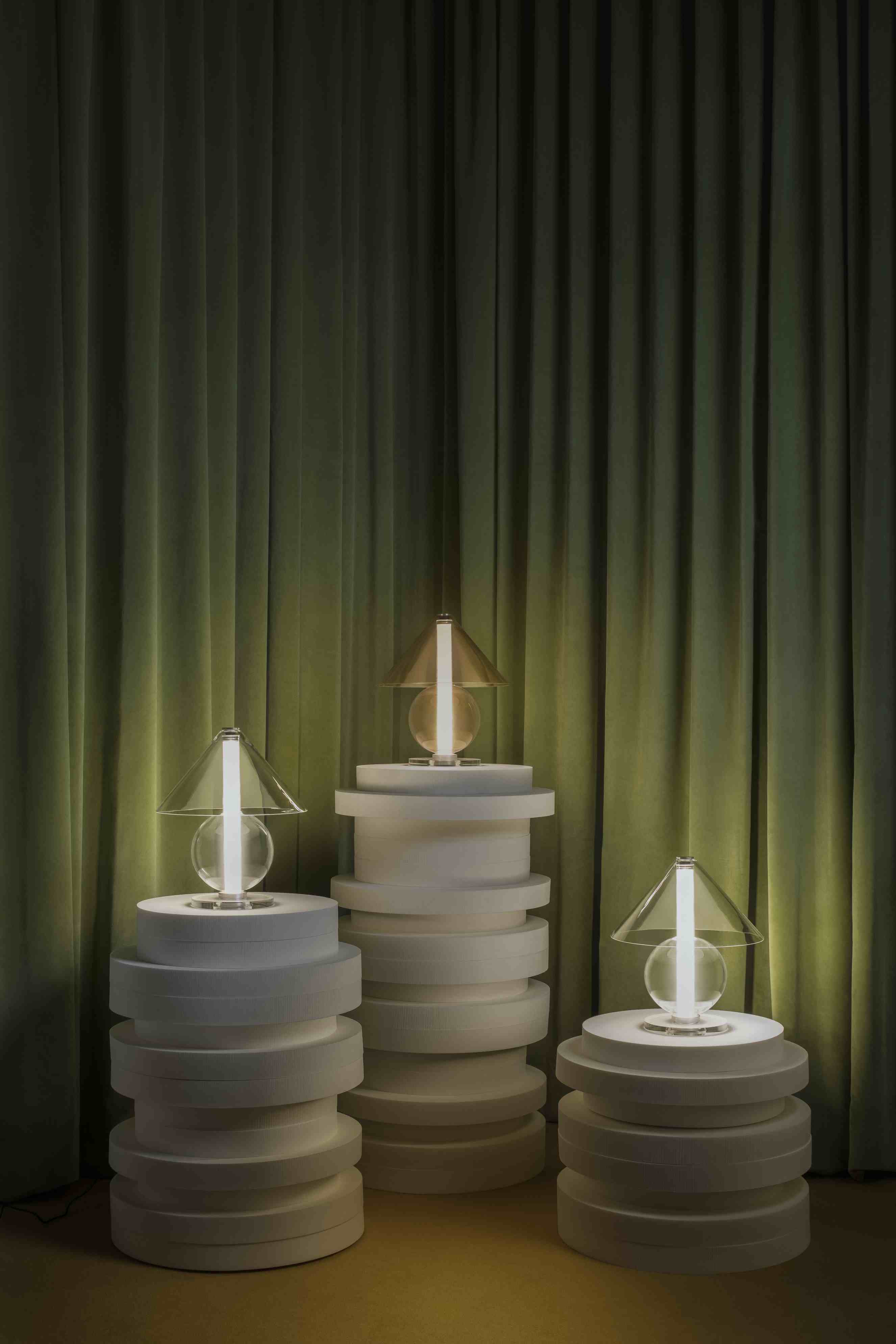 Drei Fragile Tischleuchten von Marset auf weißen Podesten vor grünem Vorhang.
