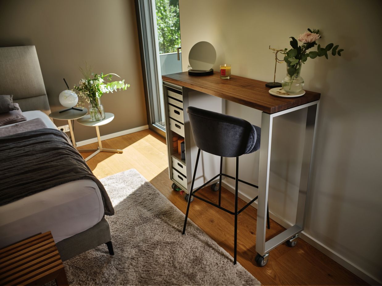 Schminktisch mit Holzplatte, Rollen und grauem Barhocker im Schlafzimmer. Spiegel, Blumen und Dekoration auf dem Tisch.