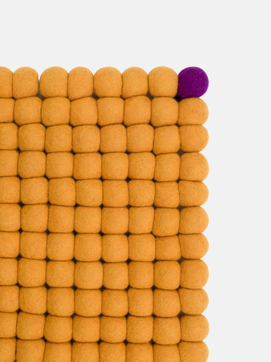 Detailaufnahme: Ockergelber Filzteppich aus vielen Filzkugeln, eine lilafarbene Kugel sticht hervor.