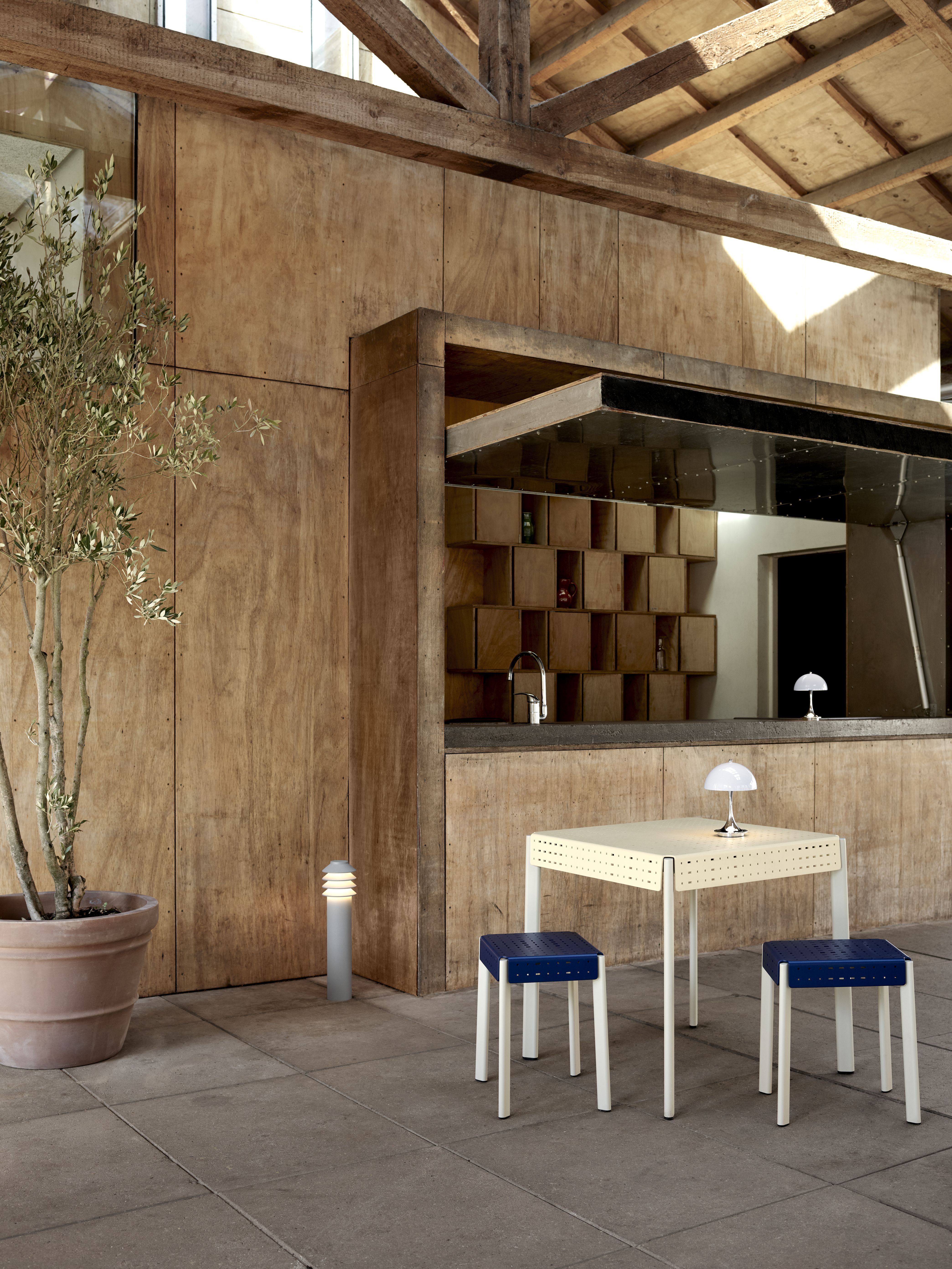 Bysted Garden Pollerleuchte von Louis Poulsen im Außenbereich neben Olivenbaum und Sitzgruppe.