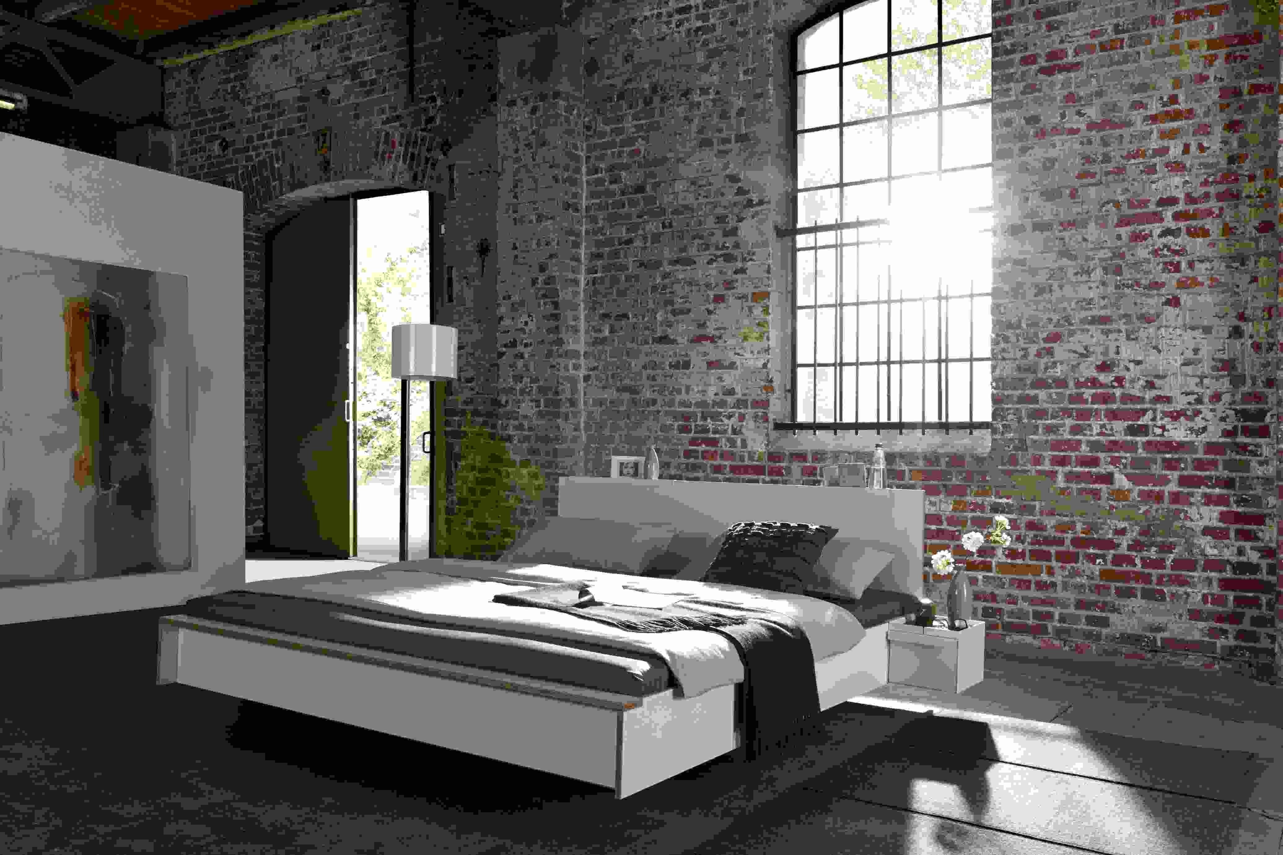 Weißes Flai Bett Kopfteil von Müller Möbelwerkstätten im Schlafzimmer mit Backsteinwand und großem Fenster.