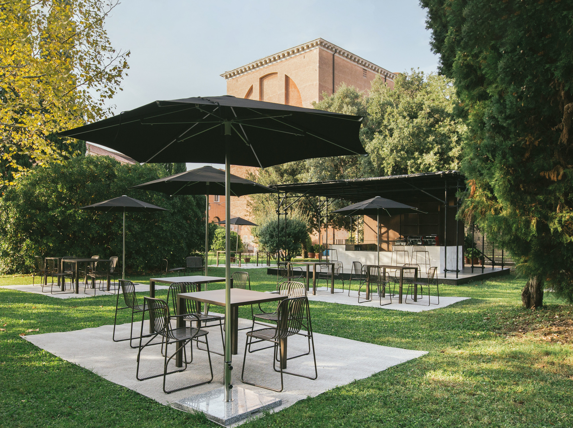 Riviera Armlehnstuhl von Emu: Schwarzer Metallstuhl im Außenbereich mit Tischen und Sonnenschirmen auf Rasen.