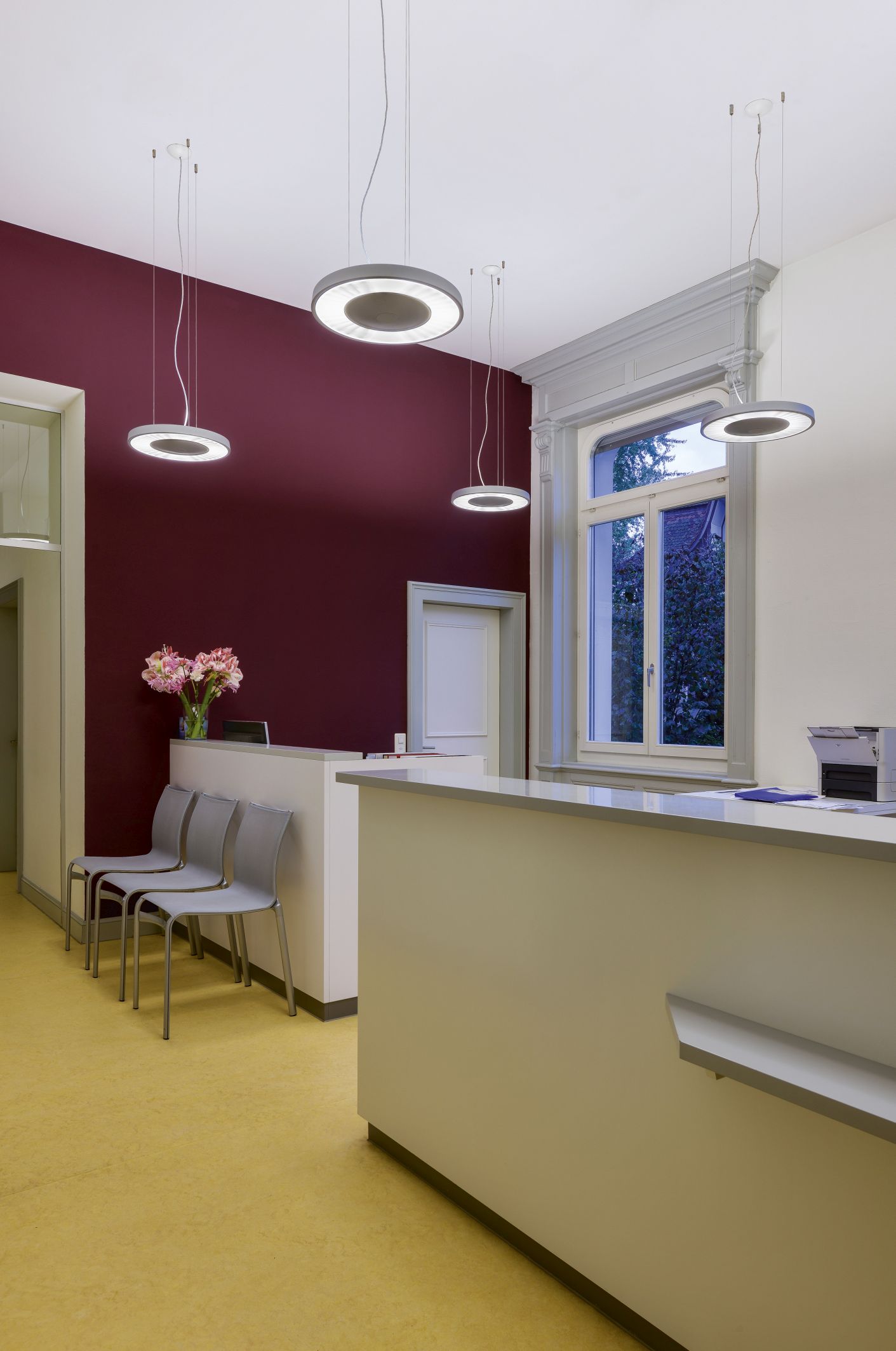 Helle Büroraum-Szene mit mehreren weißen Belux Disk Pendelleuchten 3000K über einem Empfangsbereich.