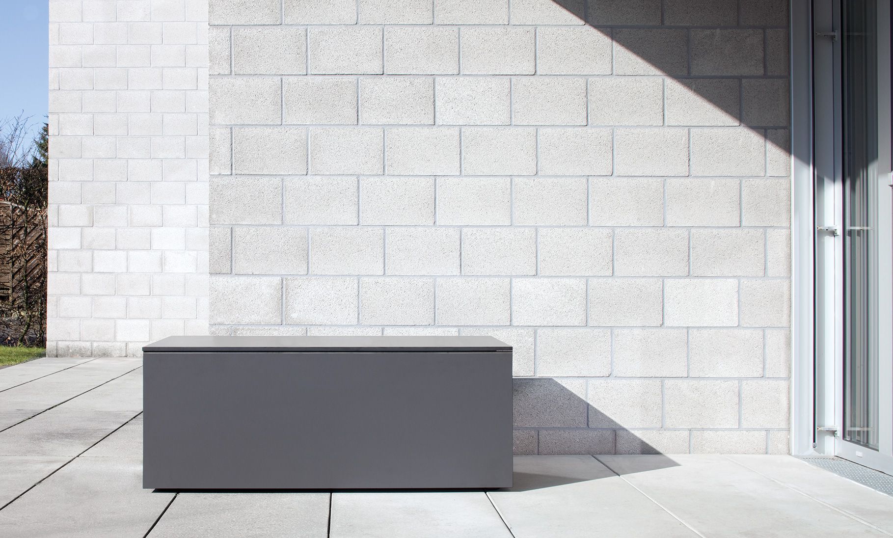 Anthrazitfarbene Kissentruhe von Conmoto, Modell EL Pecho M, auf einer Terrasse vor einer Mauer.