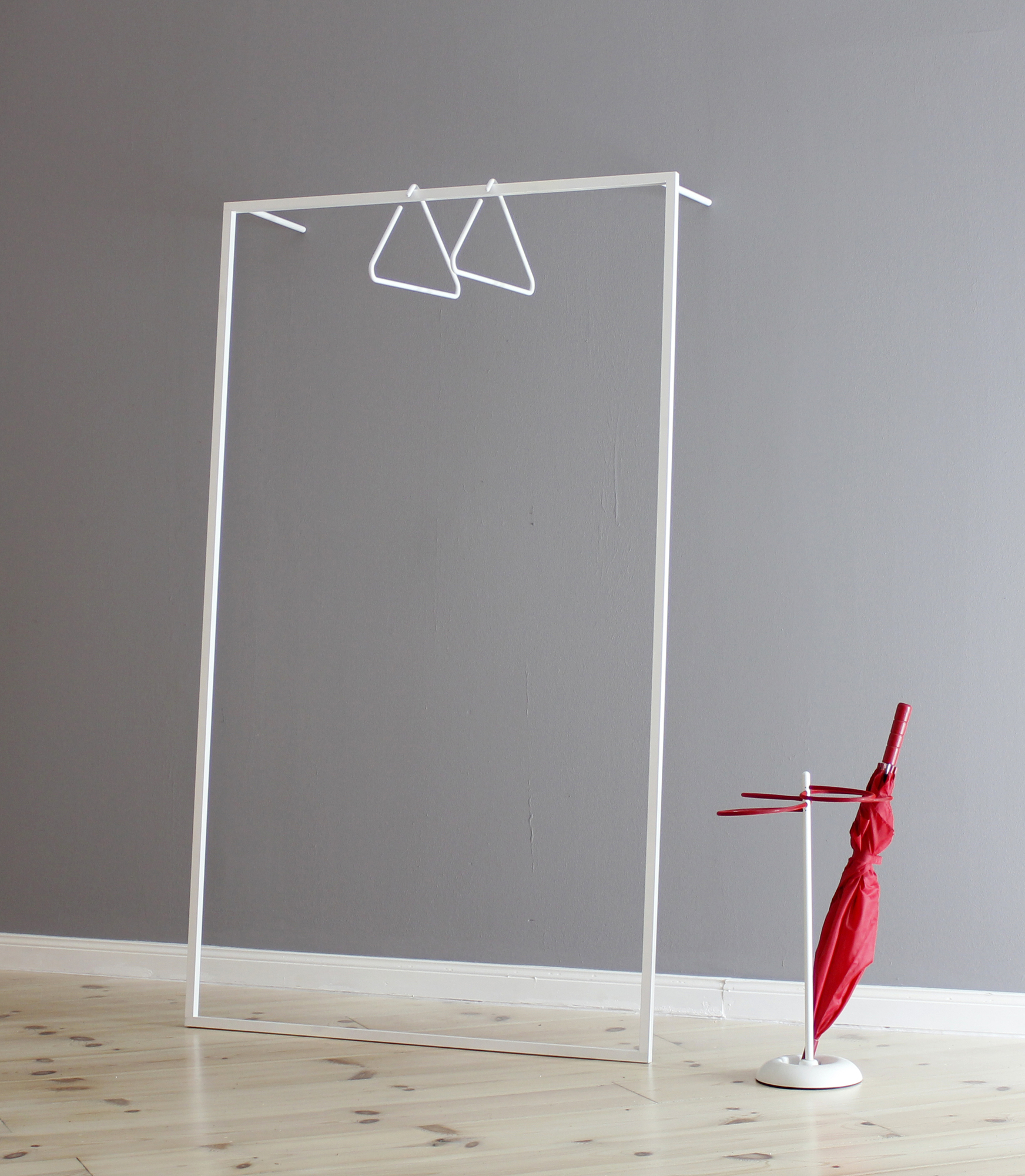 Weiße Leano Anlehngarderobe von Roomsafari mit zwei Kleiderbügeln und rotem Regenschirm vor grauer Wand.