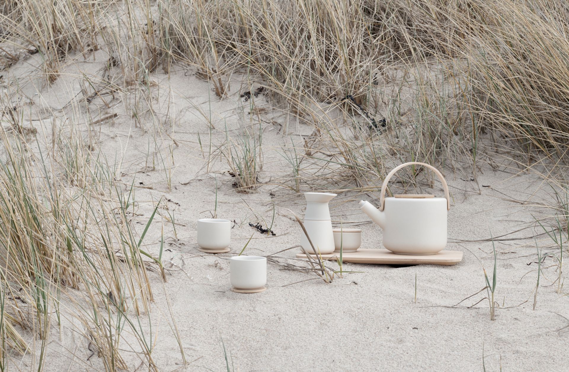 Beige Keramik Teekanne mit Tassen und Kännchen auf Holztablett im Sand, minimalistisches Teeservice.
