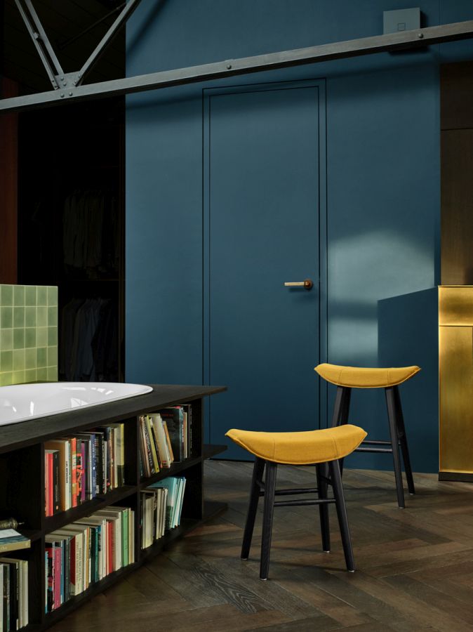 Zwei gelbe Kya Sitzhocker der Freifrau Manufaktur mit schwarzen Beinen vor blauer Wand.