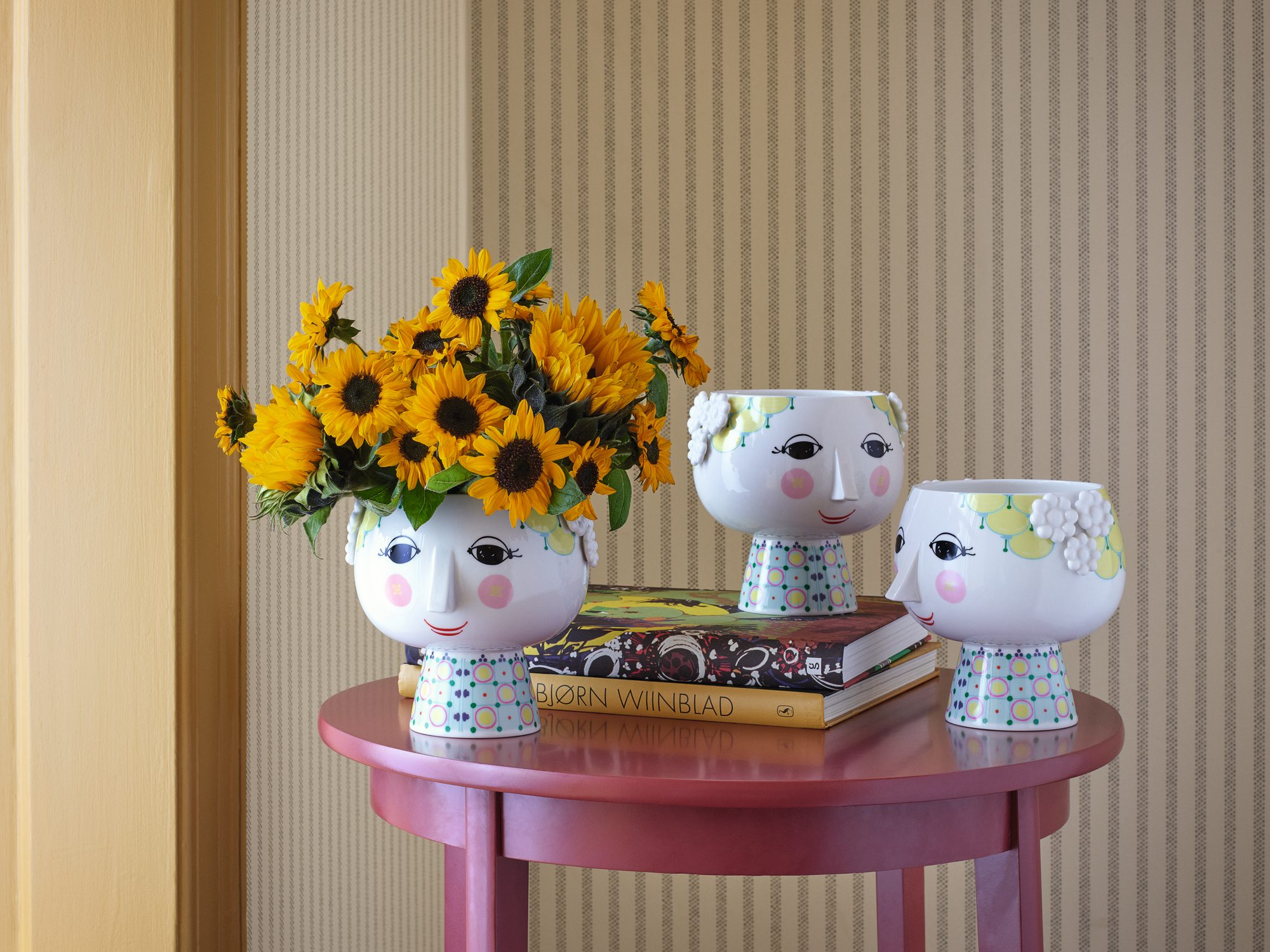 Übertopf Eva von Bjorn Wiinblad mit Sonnenblumen auf einem roten Tisch, weitere Übertöpfe im Hintergrund.