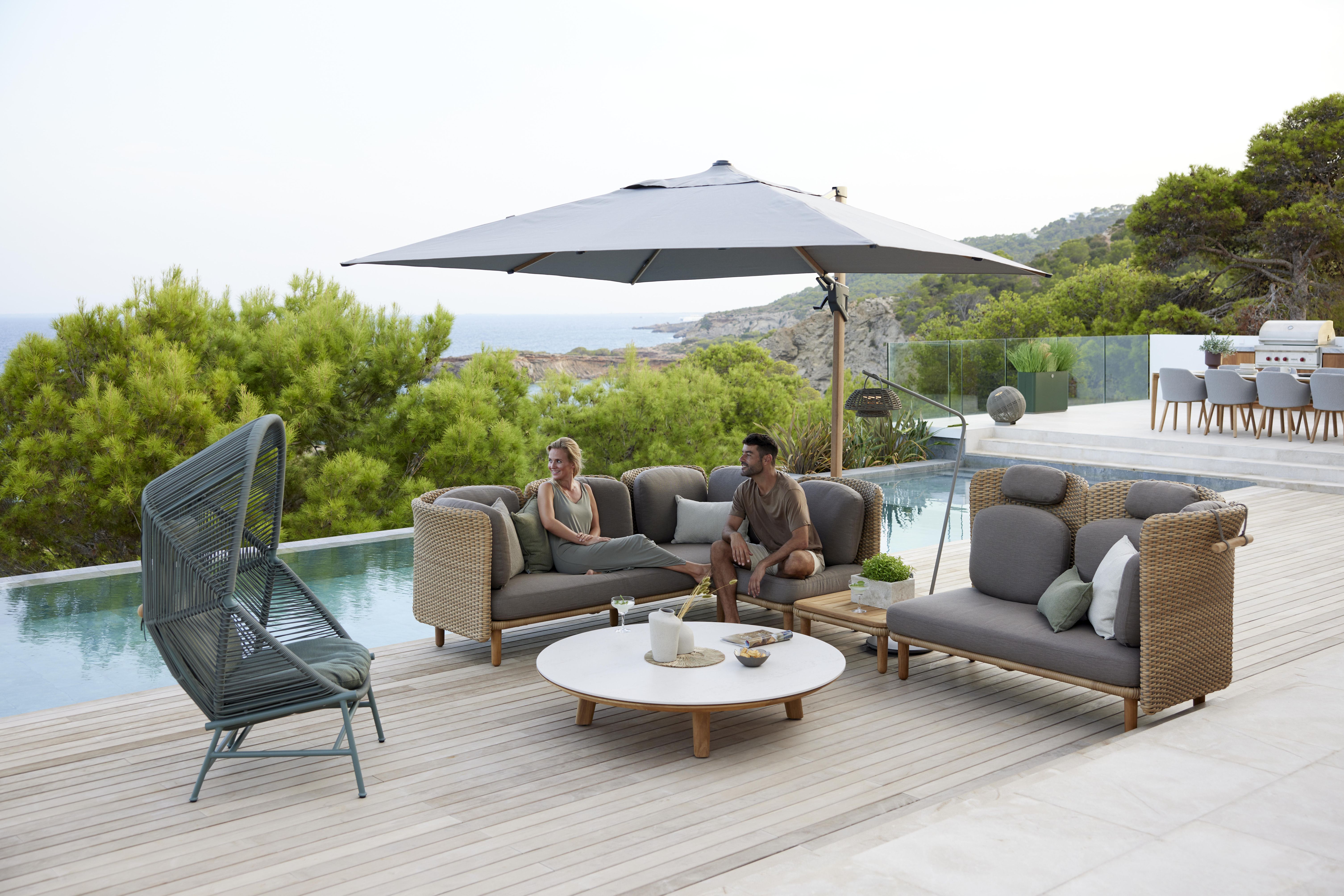 Arch Outdoor Couchtisch mit Teakplatte von Cane-Line auf einer Terrasse mit Pool und Sitzmöbeln.