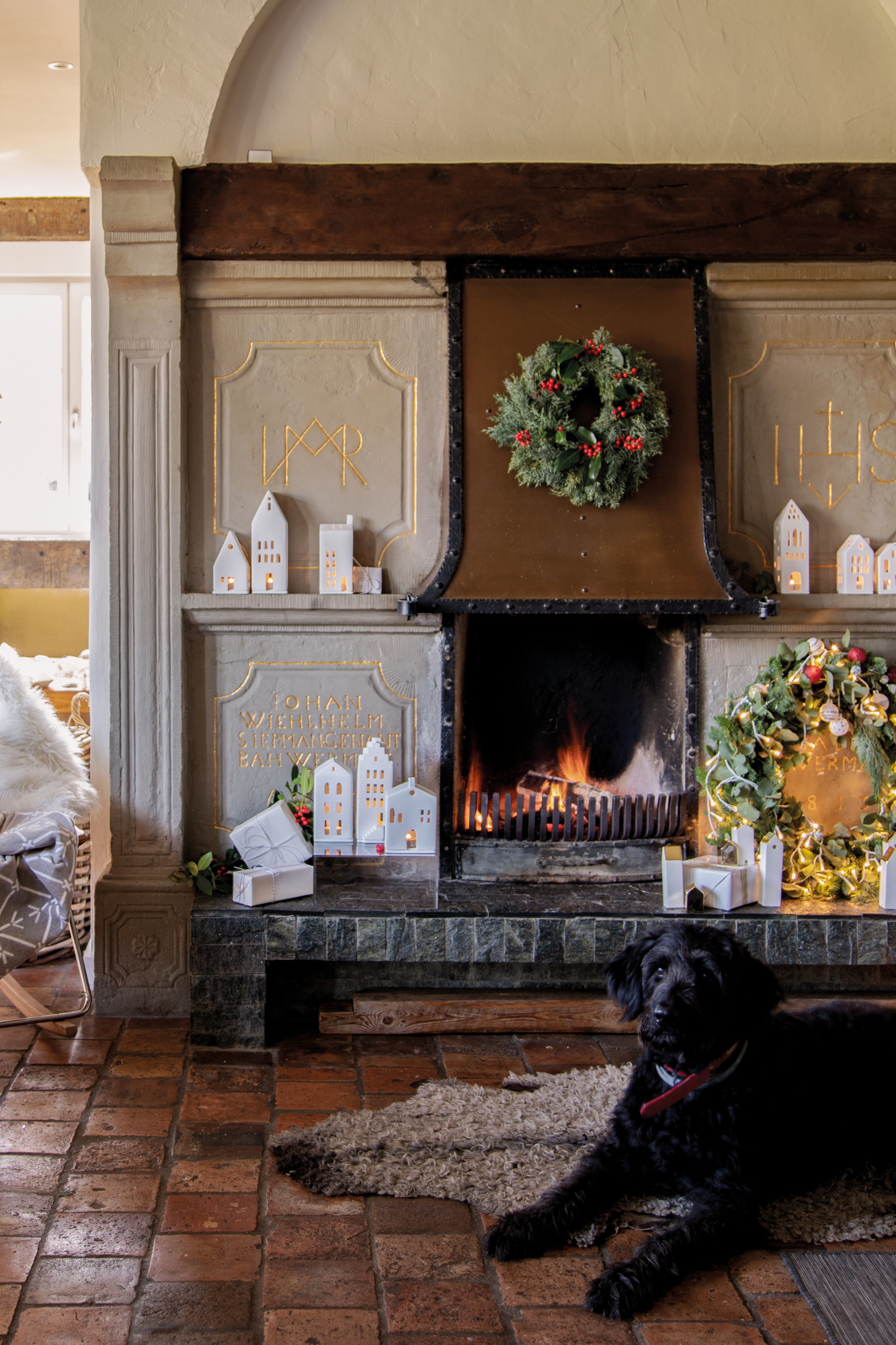 Dekorativer Kamin mit Lichthäusern, Kränzen und einem schwarzen Hund im Vordergrund. Zuhause Lichthaus als stimmungsvolle Weihnachtsdekoration.