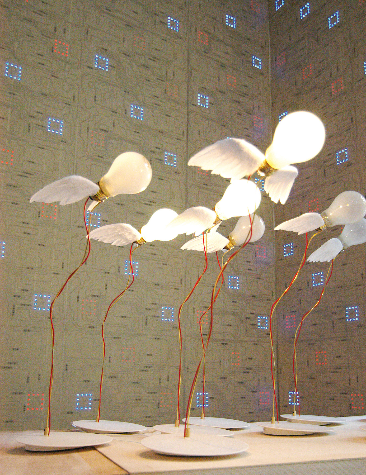 Mehrere Lucellino Tischleuchten von Ingo Maurer mit Flügeln und rotem Kabel auf weißen Sockeln.