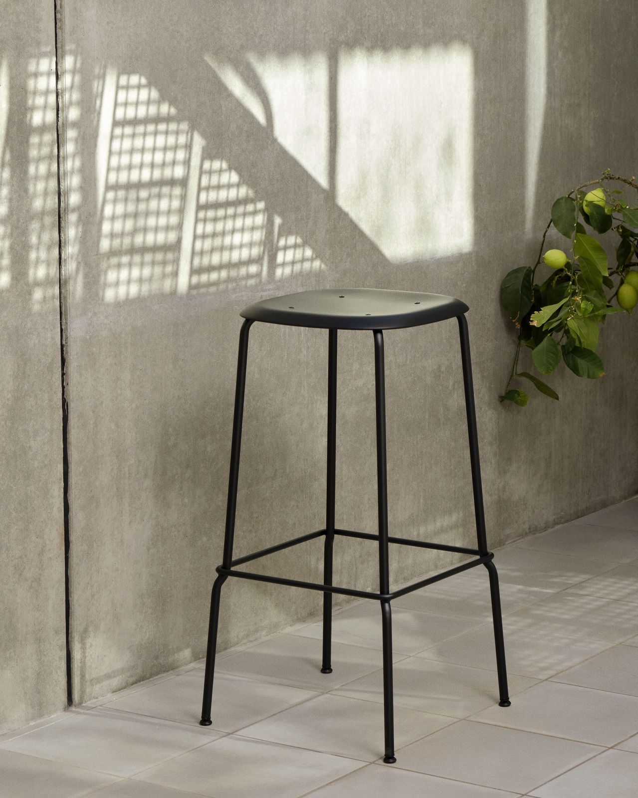 Schwarzer Hay Soft Edge Hocker 85 vor grauer Wand mit Pflanzen und Lichteffekten.