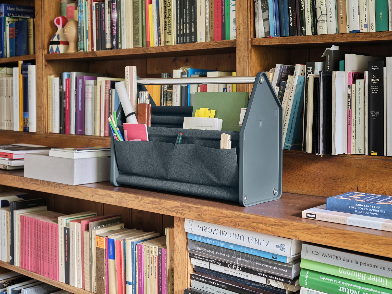 Dunkelgraue Locker Box Aufbewahrungsbox auf Bücherregal, gefüllt mit Stiften, Notizblöcken und Bürobedarf.