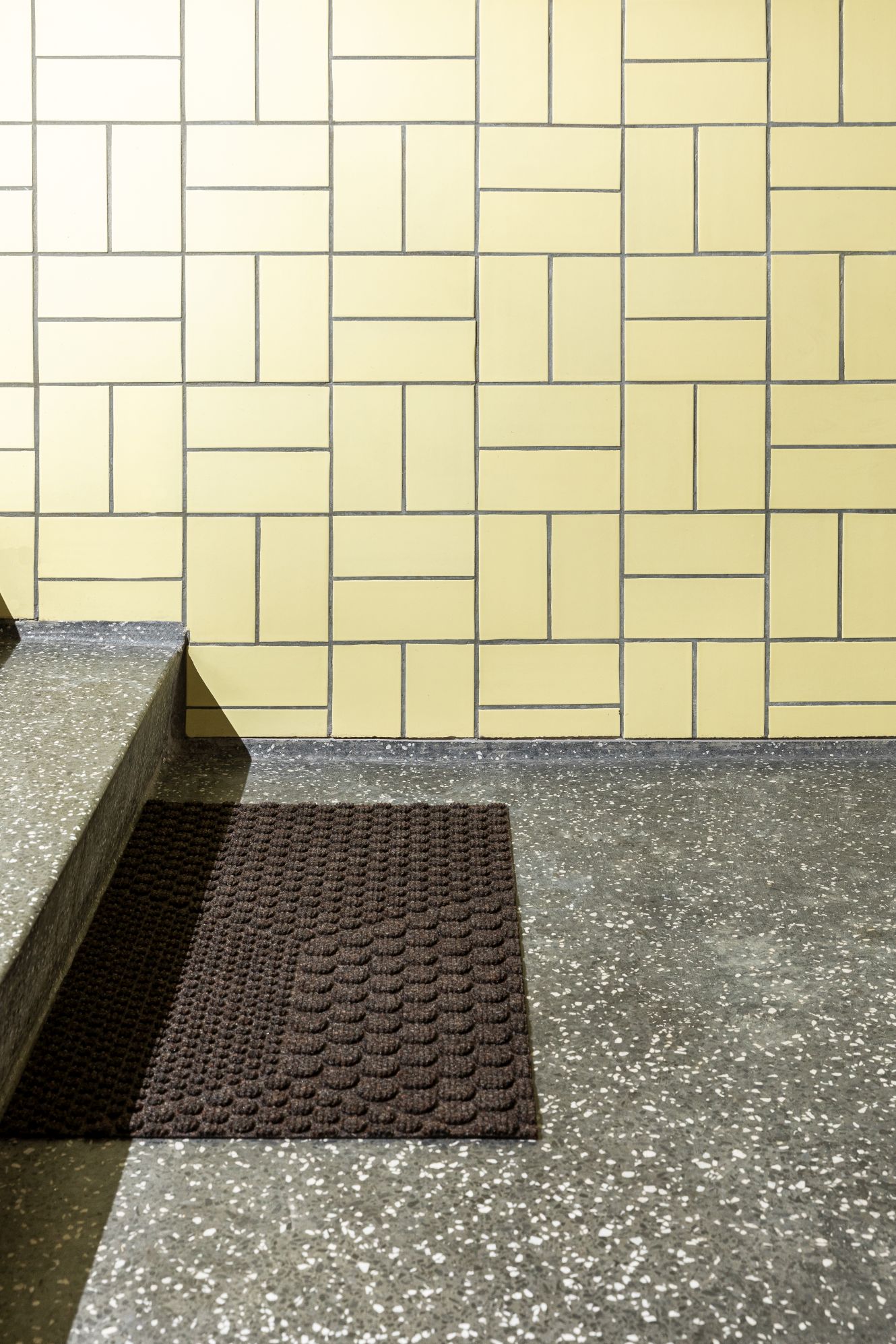 Braune Hex Fußmatte Soil von Heymat vor gelber Kachelwand und Steintreppe. Robuste Schmutzfangmatte.