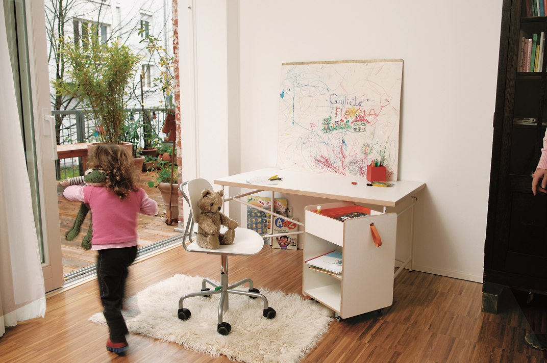 Kinderzimmer mit Kind, Schreibtisch, Stuhl und Richard Lampert Container Fixx zur Aufbewahrung von Spielsachen.