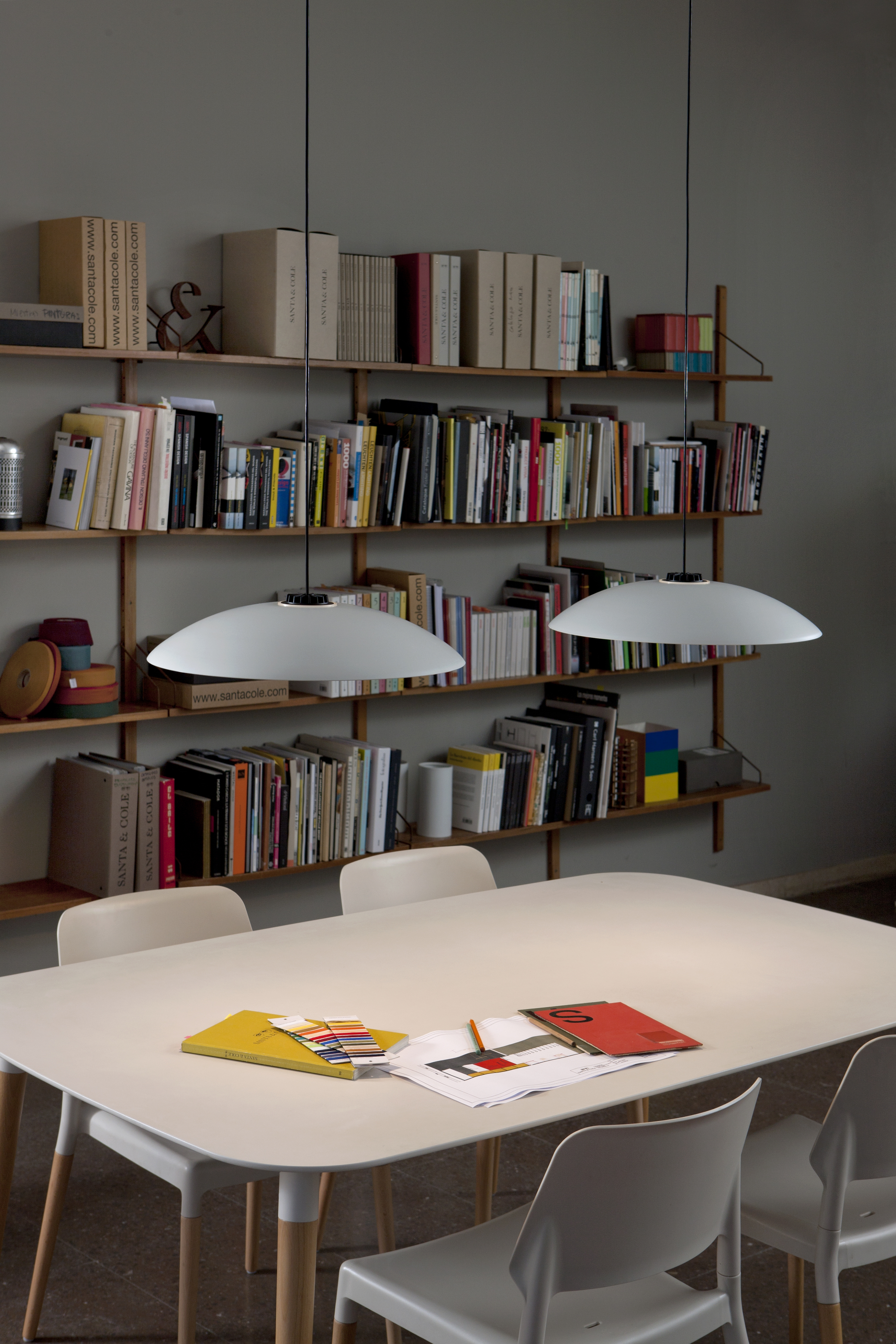 Heller Belloch Esstisch von Santa & Cole mit Stühlen und Pendelleuchten vor Bücherregal.