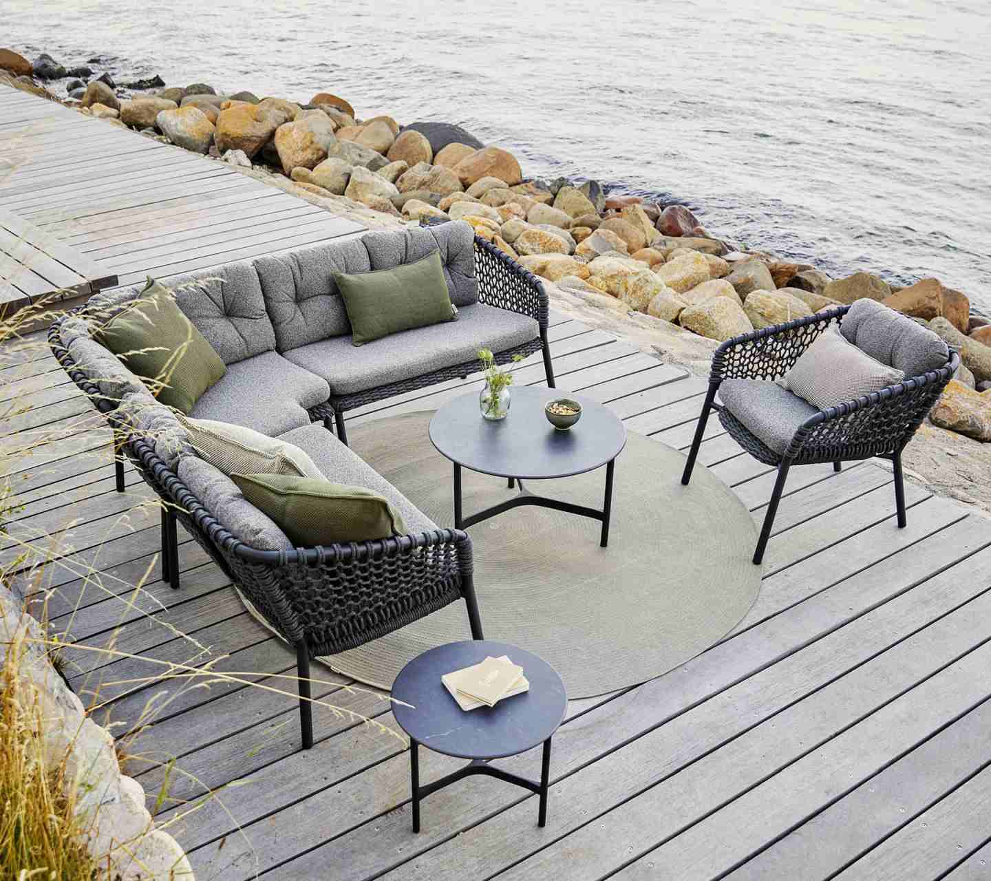 Ocean Loungesessel von Cane-Line: Outdoor-Sitzgruppe mit dunkelgrauen Kissen auf Holzterrasse am Meer.
