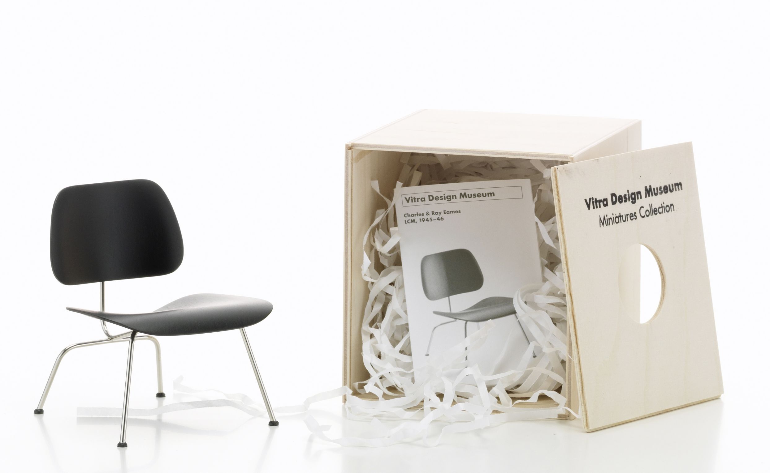 Schwarzer Vitra LCM Miniatur Stuhl [1945] mit Chromgestell neben geöffneter Holzbox mit Papierwolle.