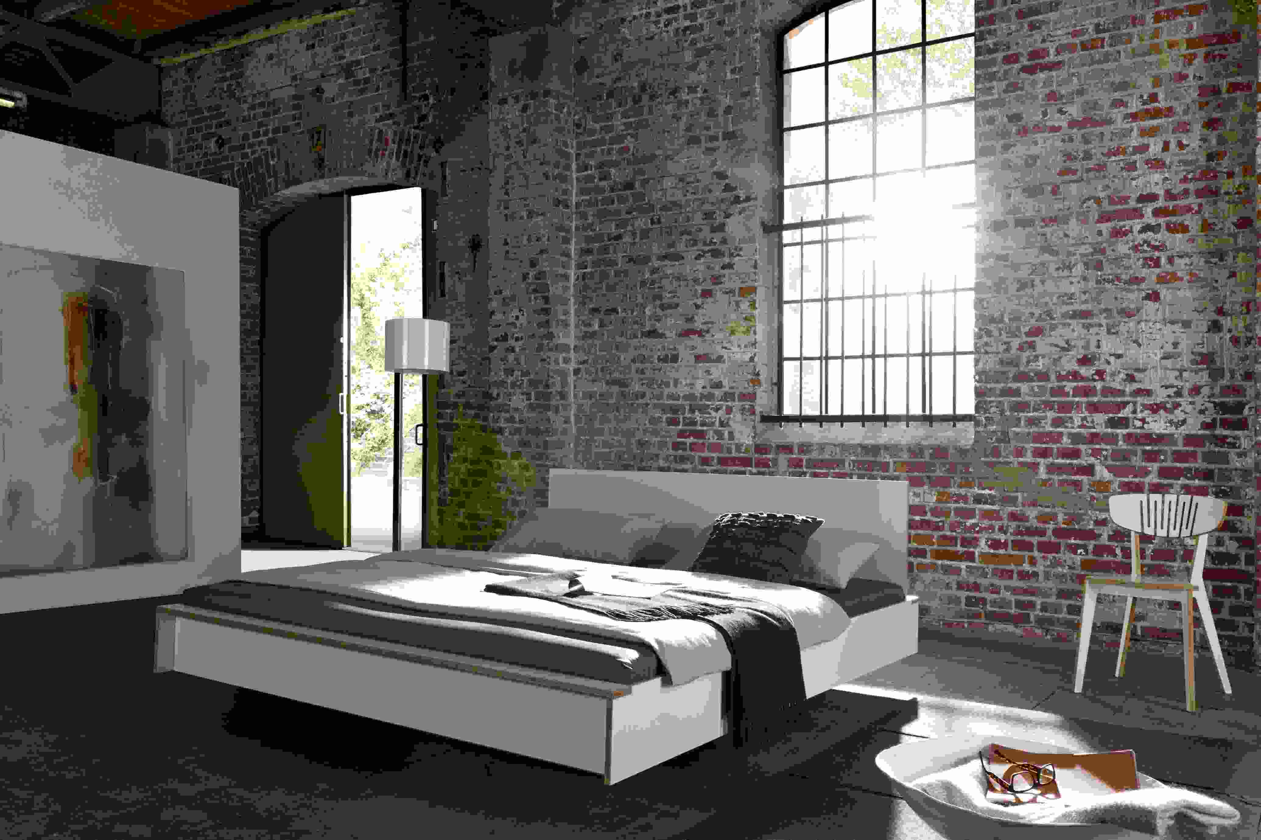 Weißes Flai Bett von Müller Möbelwerkstätten in modernem Loft mit Backsteinwand und Tageslicht.