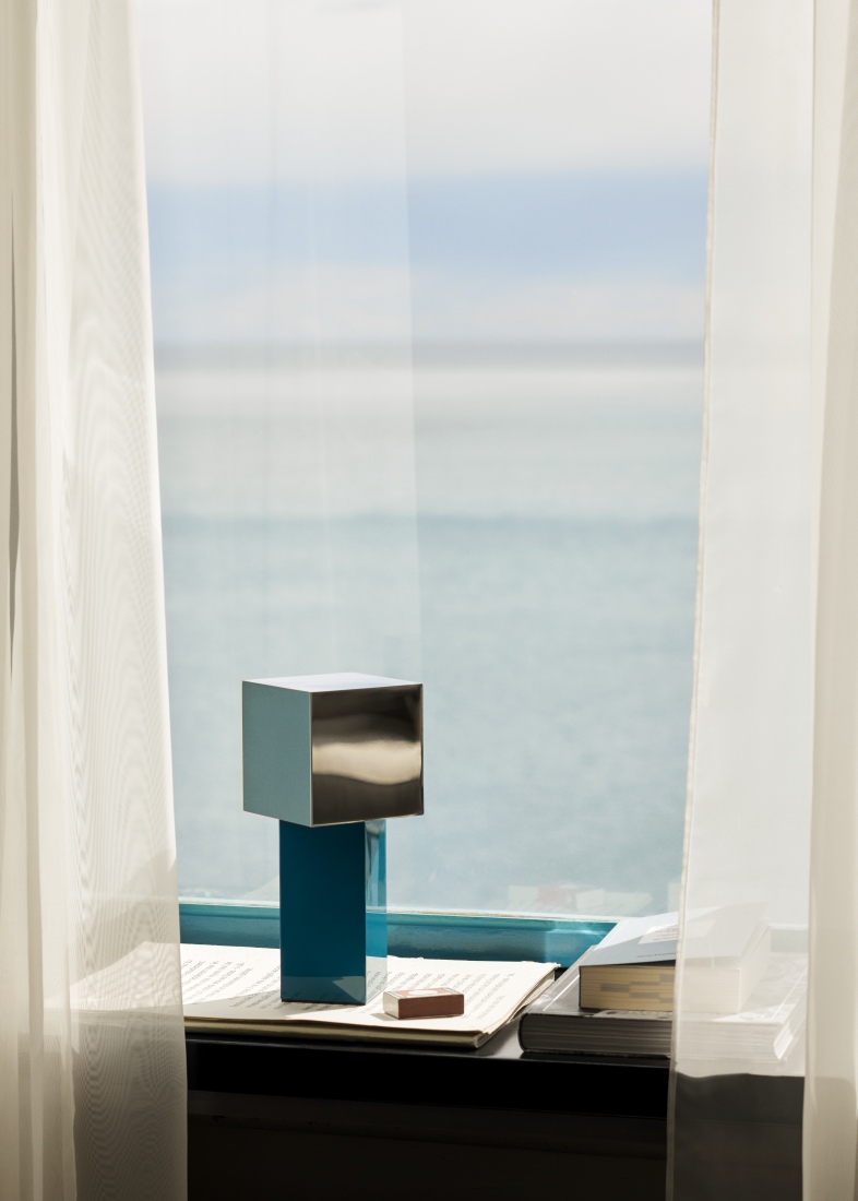 Pivot ATD7 Tischleuchte von AndTradition, blaue Akkuleuchte auf einem Tisch vor einem Fenster.