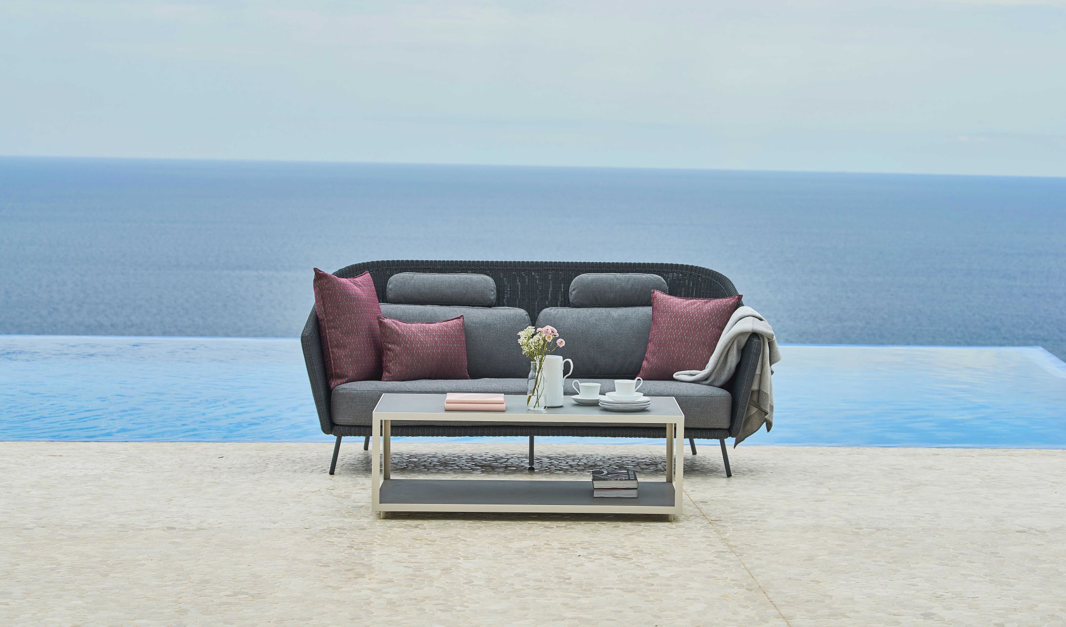 Mega Outdoor Sofa von Cane-Line mit Kissen und Tisch vor Pool und Meer.