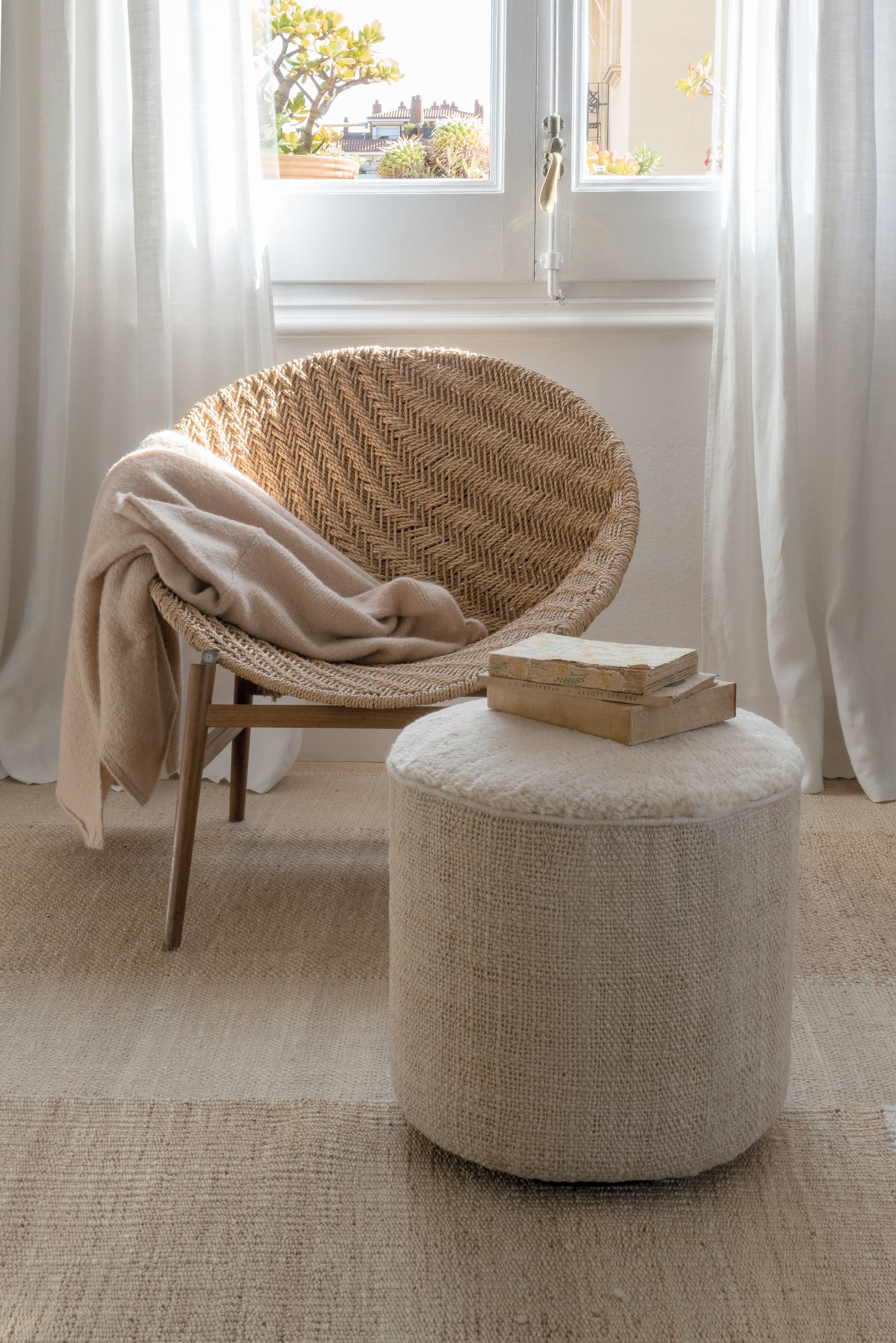 Nanimarquina Tres Persian Pouf aus Wolle in Beige, neben Rattansessel mit Decke.