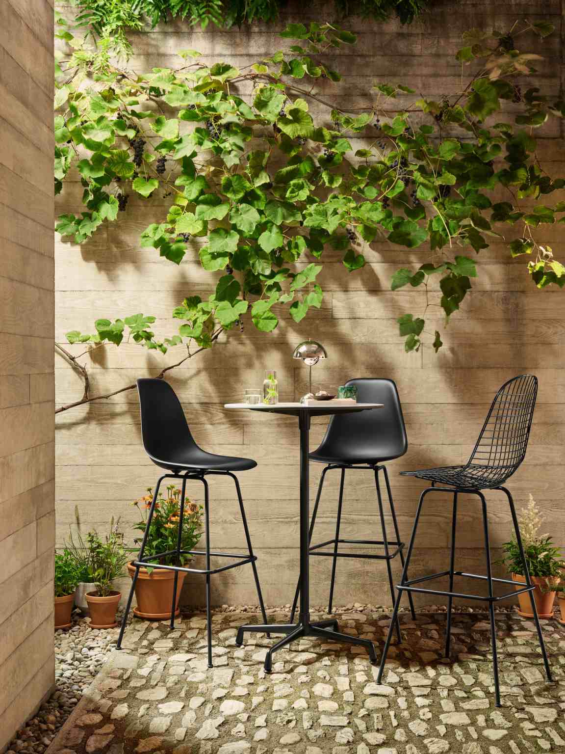 Drei schwarze Barhocker, darunter ein Eames Plastic Bar Stool, an einem Bistrotisch im Garten.