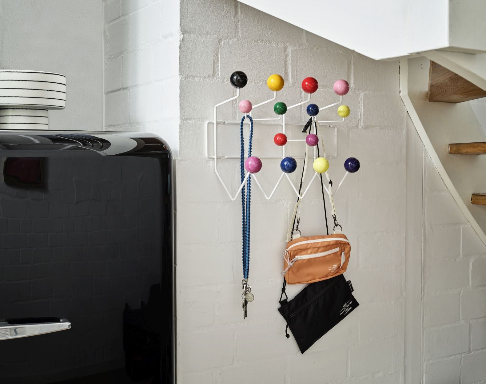 Weiße Wandgarderobe mit bunten Holzkugeln, Taschen und Schlüsselanhänger an einer weißen Ziegelwand neben einem schwarzen Kühlschrank.