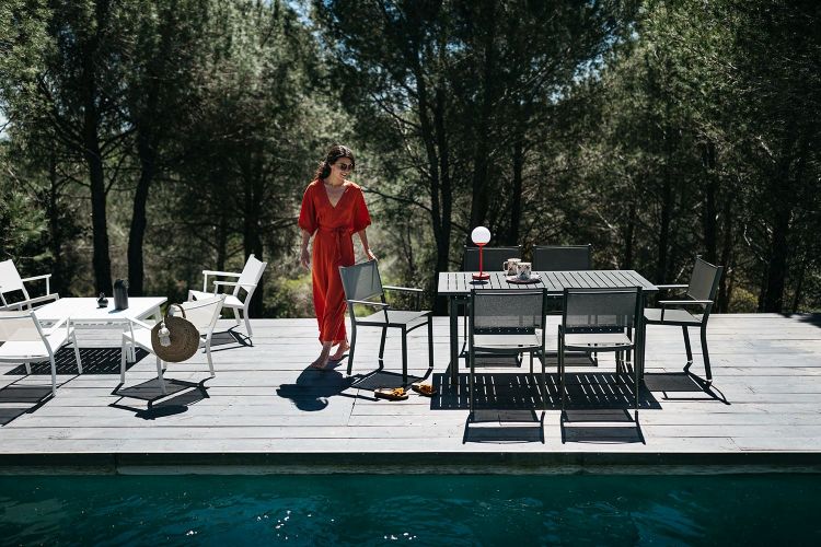 Costa Tisch von Fermob: Grauer Gartentisch mit Stühlen auf Holzterrasse am Pool, umgeben von Bäumen.